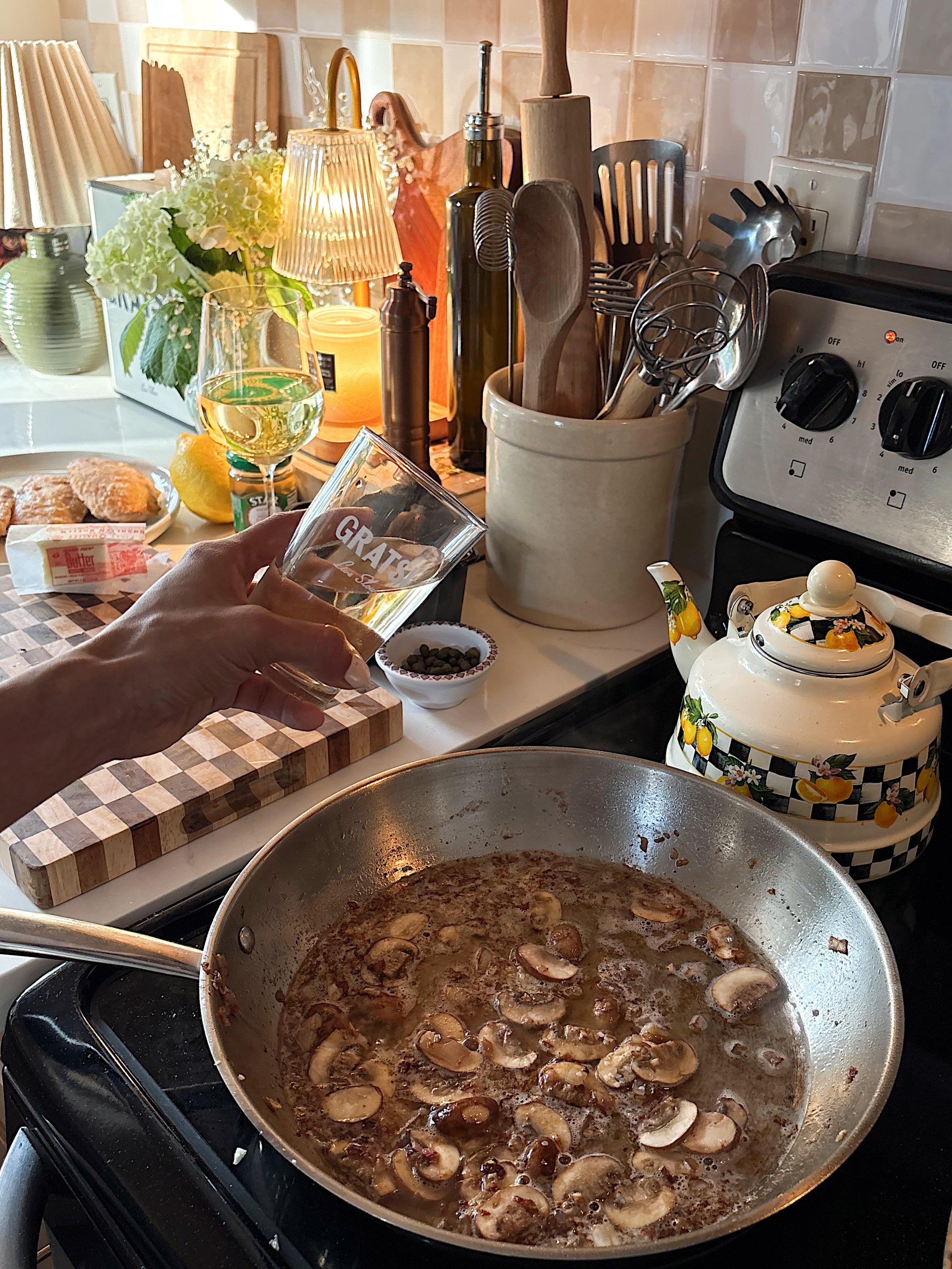 A hand pours white wine labeled GRAPE into a pan of sautéing mushrooms on a stove. Kitchen utensils, a teapot, and cutting board with food ingredients are visible in a cozy, warmly lit kitchen.
