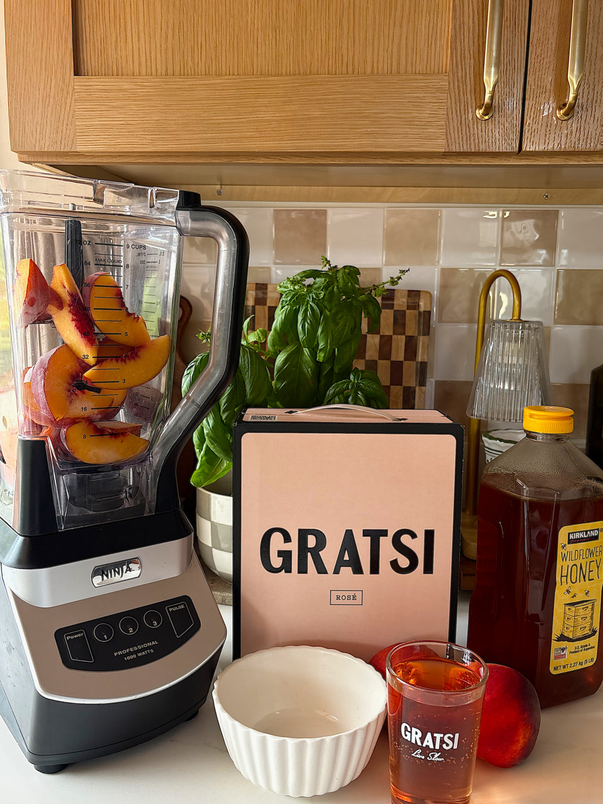 A blender with peach slices, a box labeled GRATSI, a jar of honey, a glass of juice, a peach, a bowl, and fresh basil sit on a kitchen counter with wooden cabinets and a tiled backsplash.
