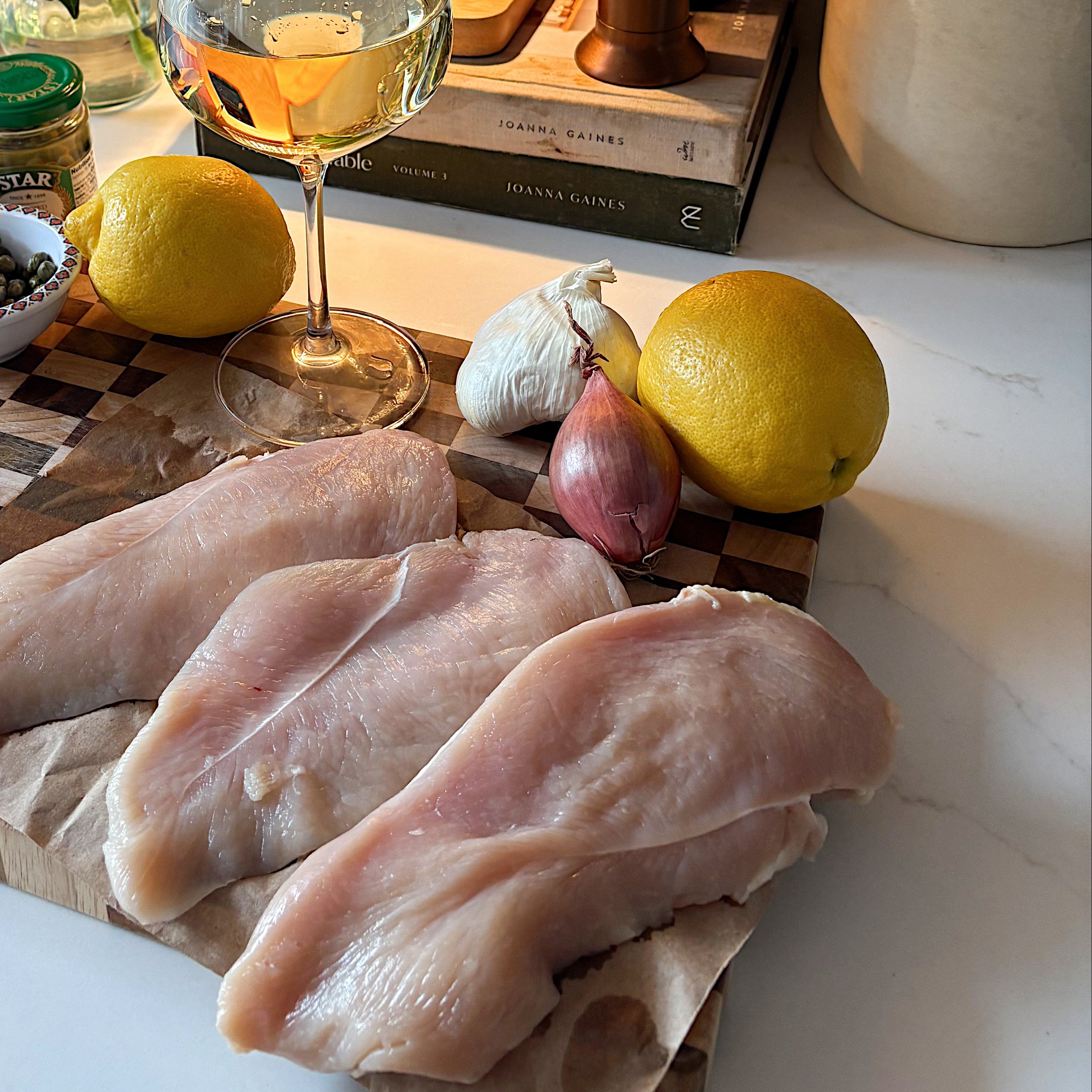 Raw chicken breasts, lemons, garlic, shallot, capers, grated cheese, flour, and a glass of white wine are arranged on a cutting board in a cozy kitchen with warm lighting, flowers, and kitchen utensils in the background.