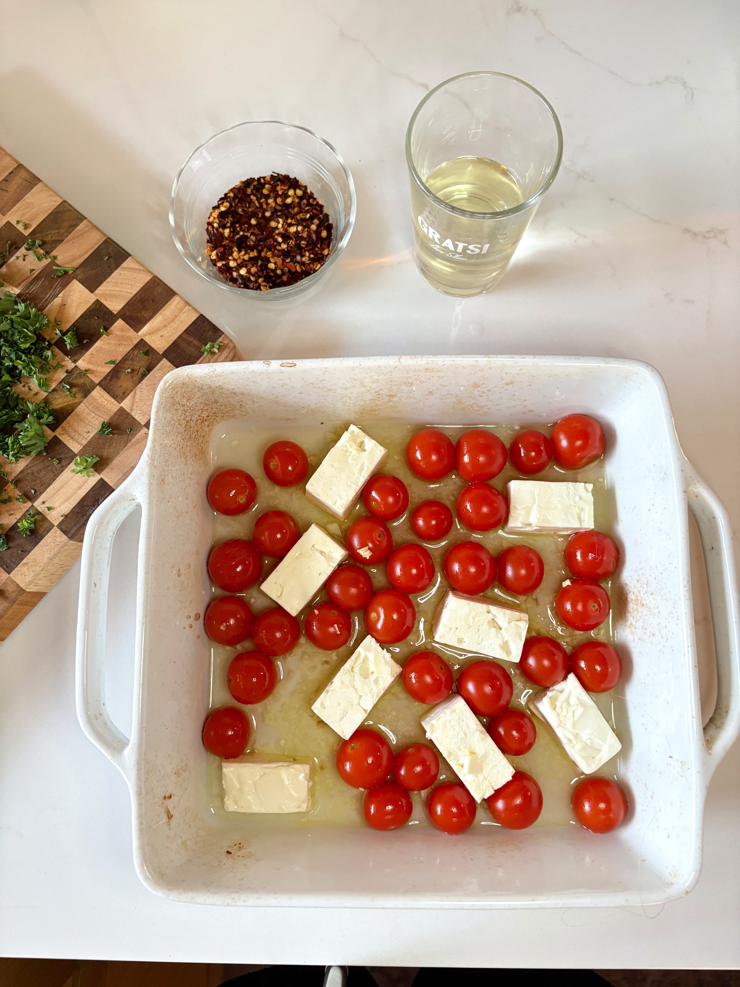 A white baking dish with cherry tomatoes and blocks of cheese in olive oil, next to a bowl of red pepper flakes, a glass of white wine, and chopped herbs on a wooden cutting board.