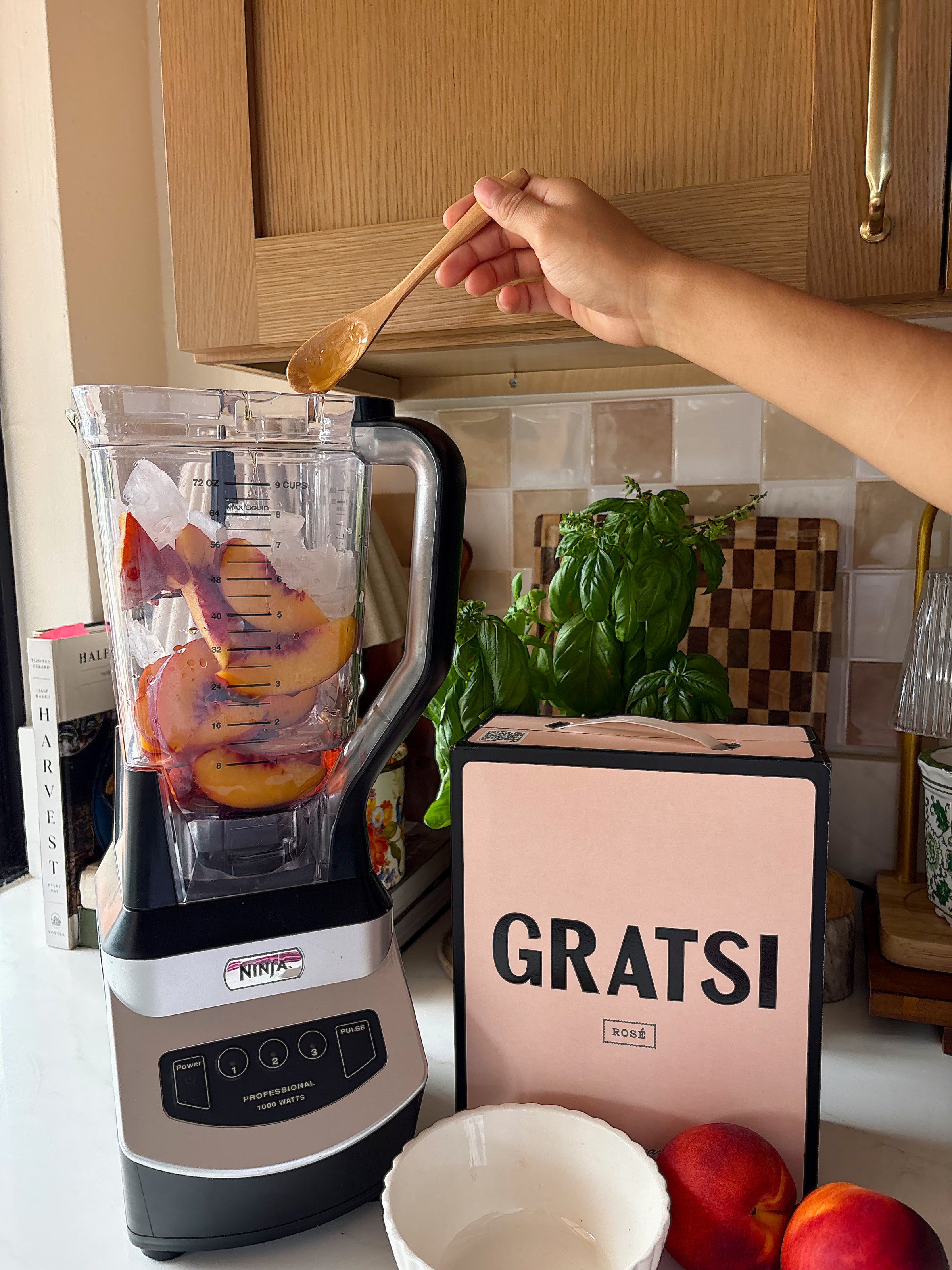 A hand holds a wooden spoon over a blender filled with ice and peach slices. Next to it are a pink GRATS! box, a white bowl, peaches, and a basil plant on a kitchen counter.