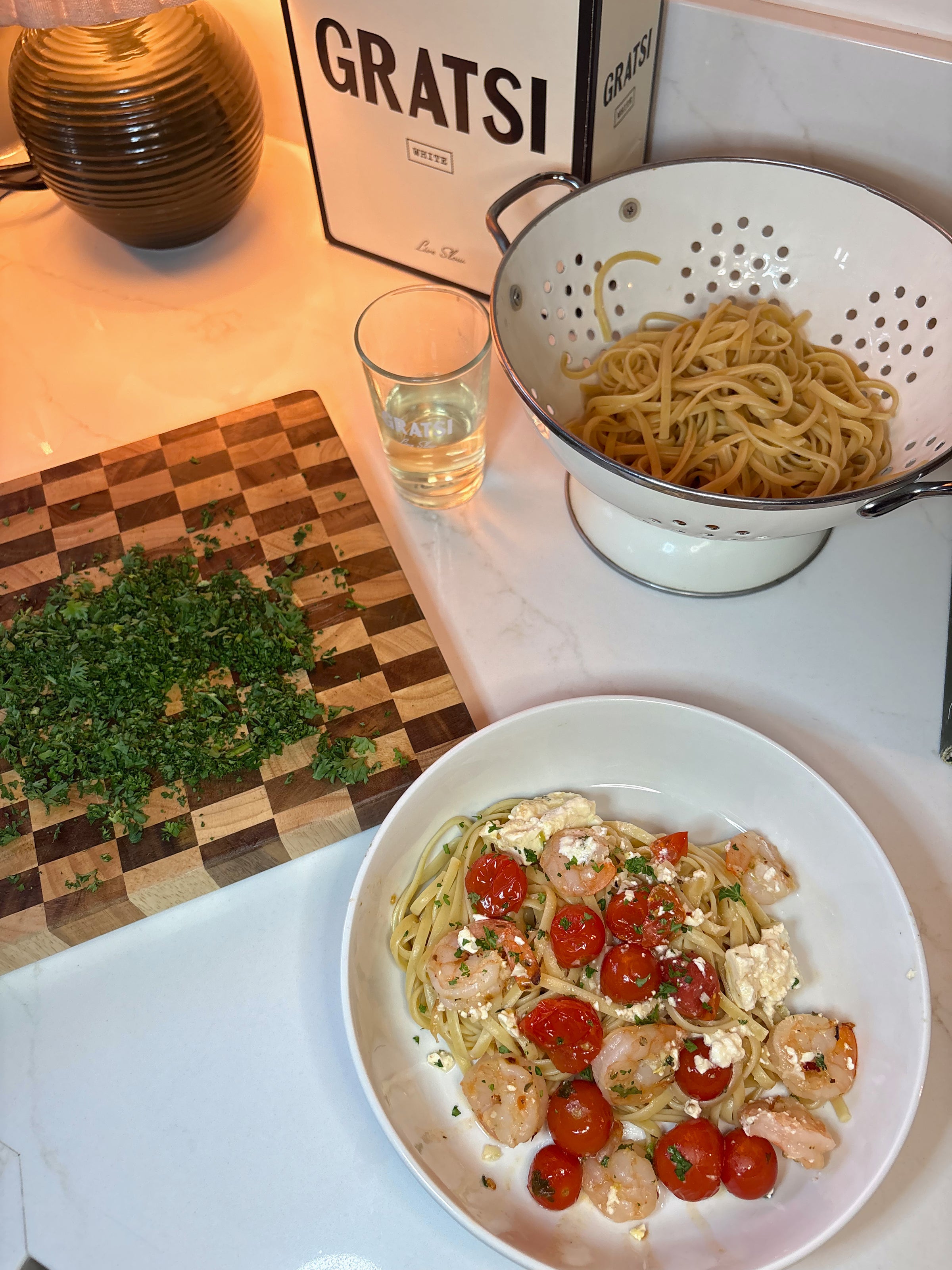 A bowl of pasta with shrimp, cherry tomatoes, and crumbled cheese is on a counter. Nearby are a cutting board with chopped herbs, a colander with plain pasta, a glass of water, and a Gratsi wine box.