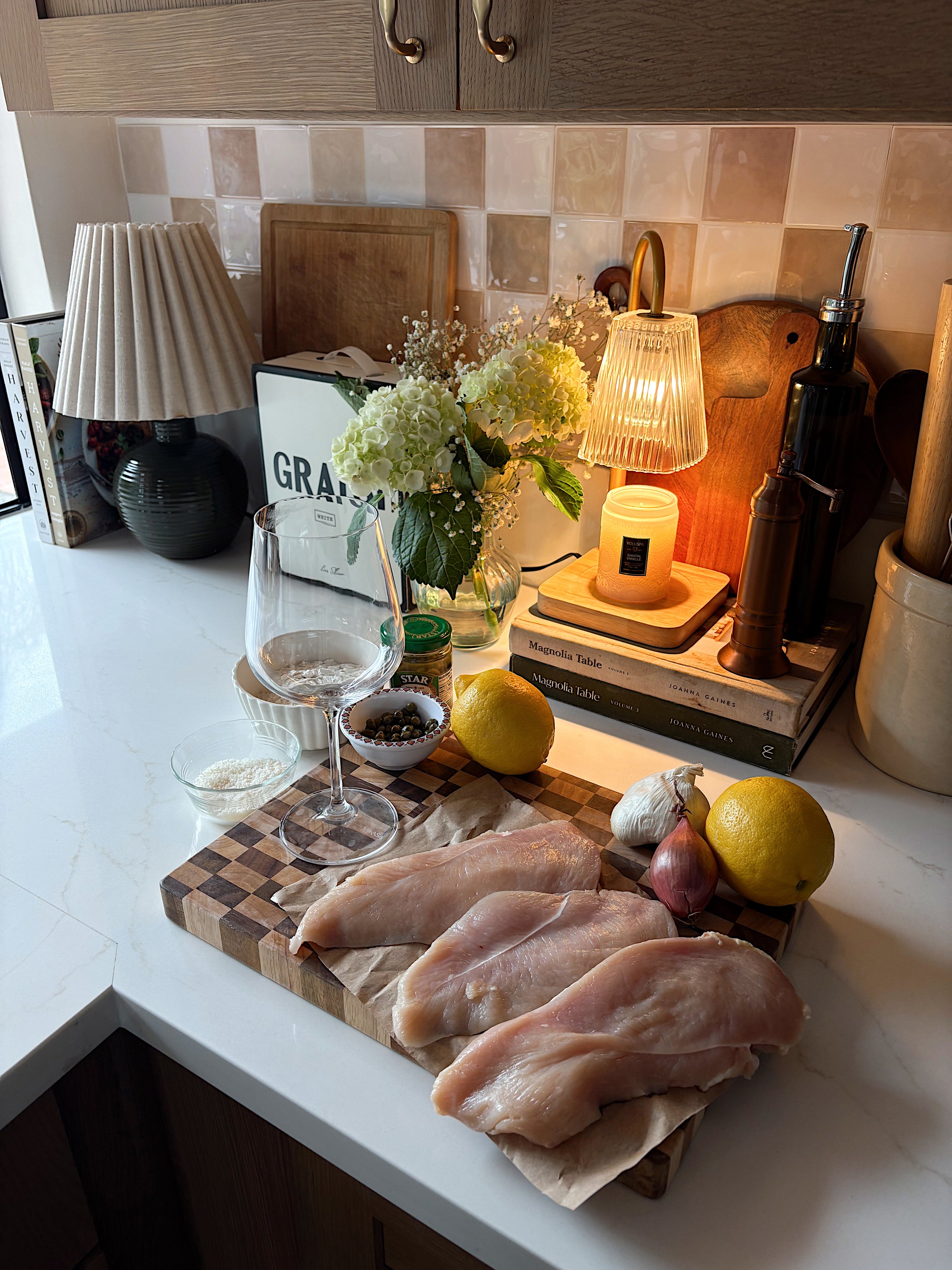 Raw chicken breasts on a wooden cutting board with lemons, shallots, garlic, wine glasses, salt, peppercorns, olive oil, and flowers on a kitchen counter lit by warm lamps.