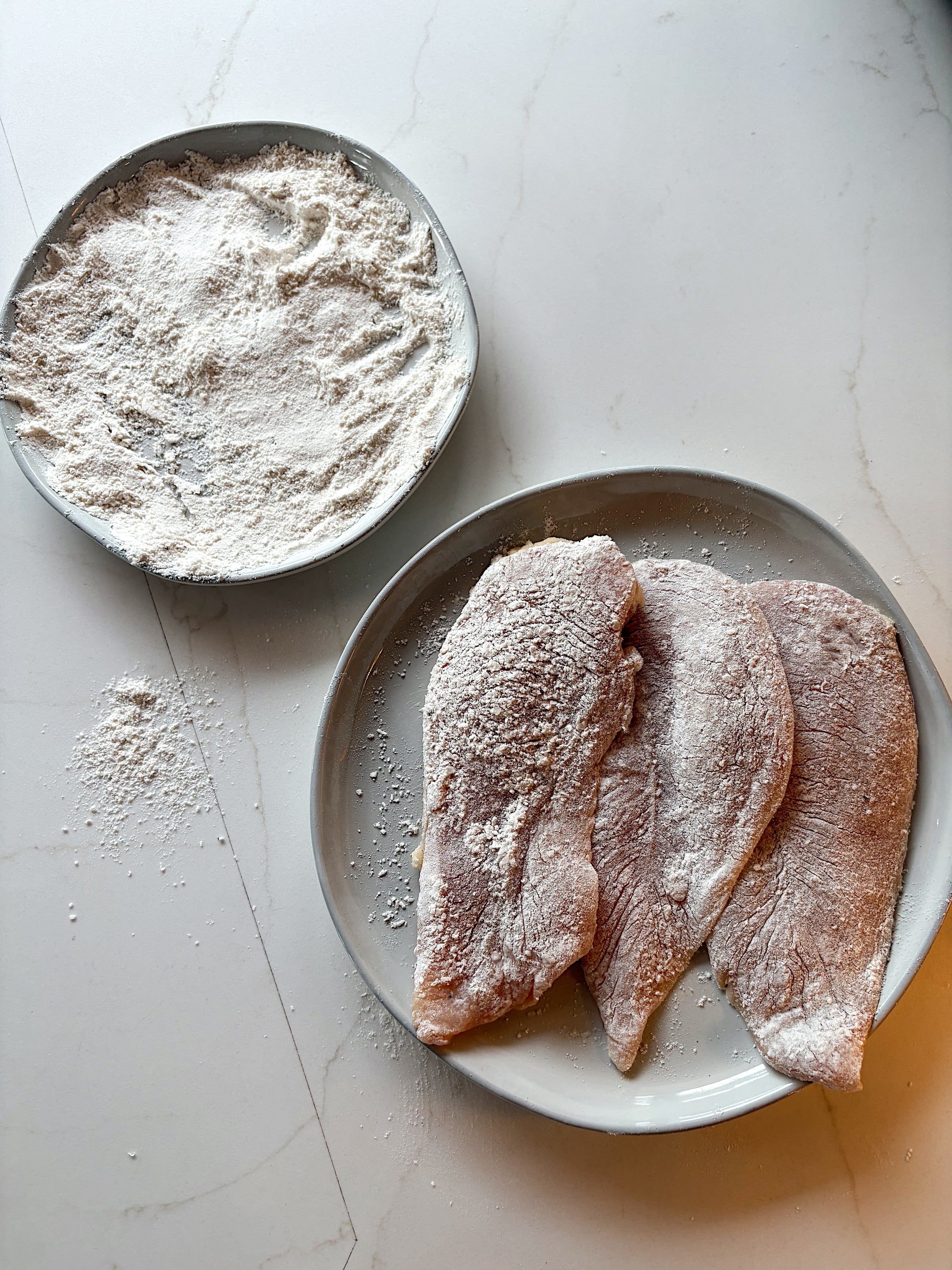 Two plates on a countertop: one with a pile of seasoned flour, the other with three raw chicken cutlets coated lightly in flour, with some flour spilled onto the counter.