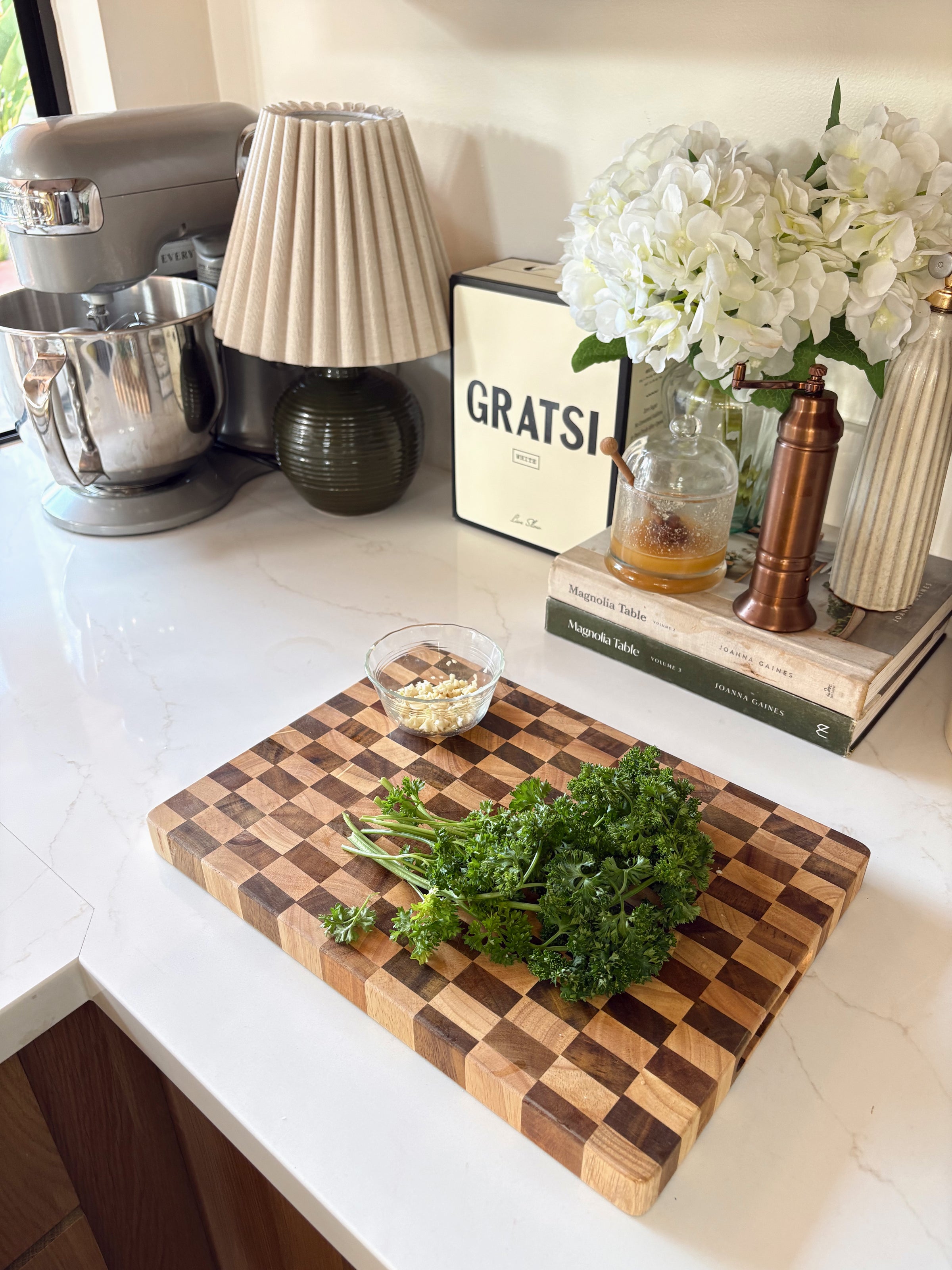 A checkered wooden cutting board with fresh parsley and a small bowl of minced garlic sits on a white kitchen counter near a mixer, lamp, books, flowers, and a bottle of olive oil.