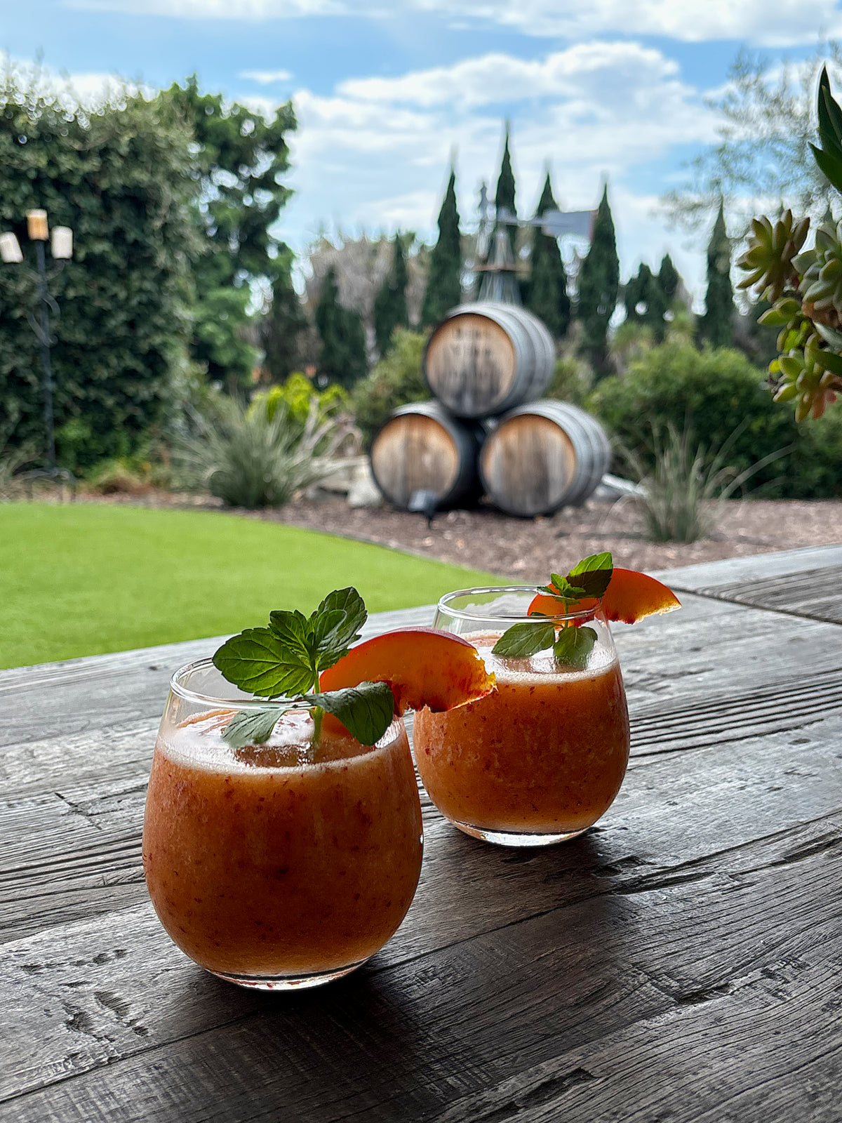 Two glasses of peach-colored drinks garnished with mint and peach slices sit on a wooden table outdoors, with stacked wine barrels and greenery visible in the background.