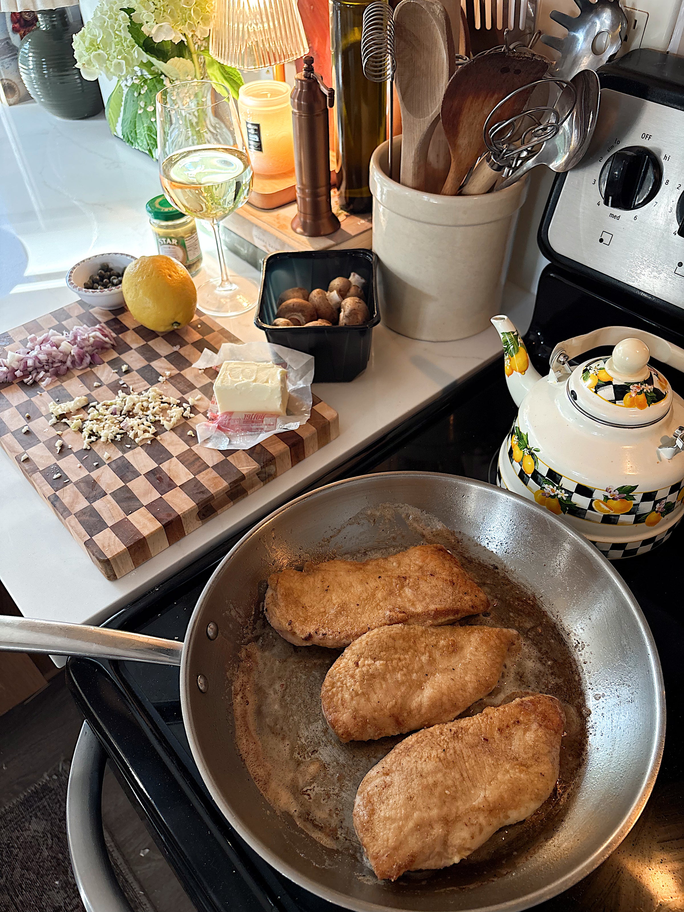 Three breaded chicken cutlets cook in a pan on a stove. Nearby are a teapot, utensils, a glass of white wine, a cutting board with chopped vegetables, mushrooms, lemon, cheese, and a jar of capers.