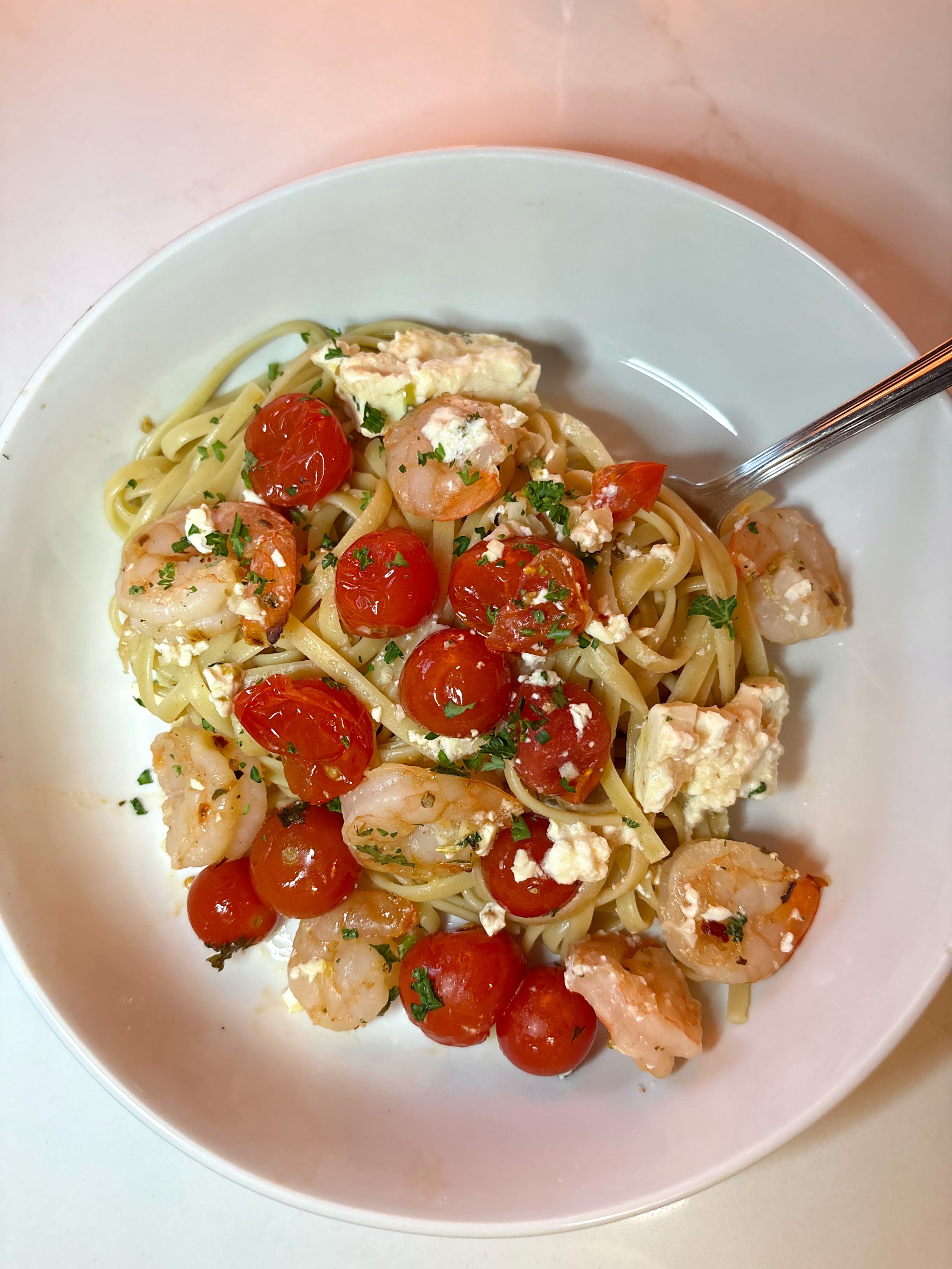 A bowl of linguine pasta topped with shrimp, cherry tomatoes, crumbled feta cheese, and chopped parsley, with a fork resting on the side.