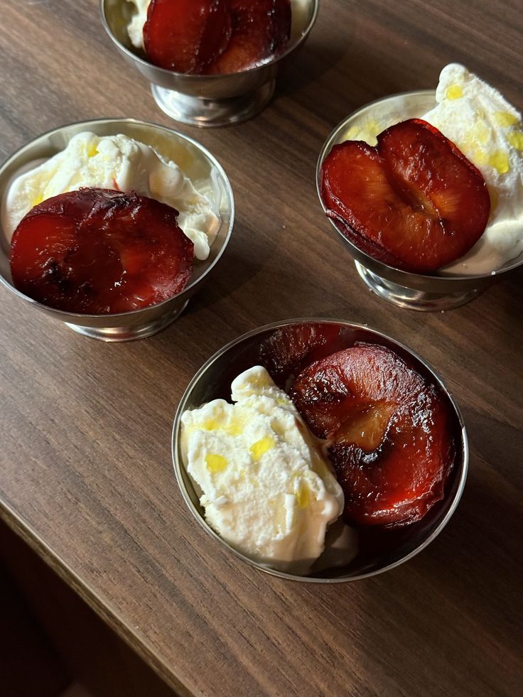 Three small metal bowls each contain a serving of vanilla ice cream topped with roasted halved plums and a drizzle of olive oil, arranged on a wooden surface.