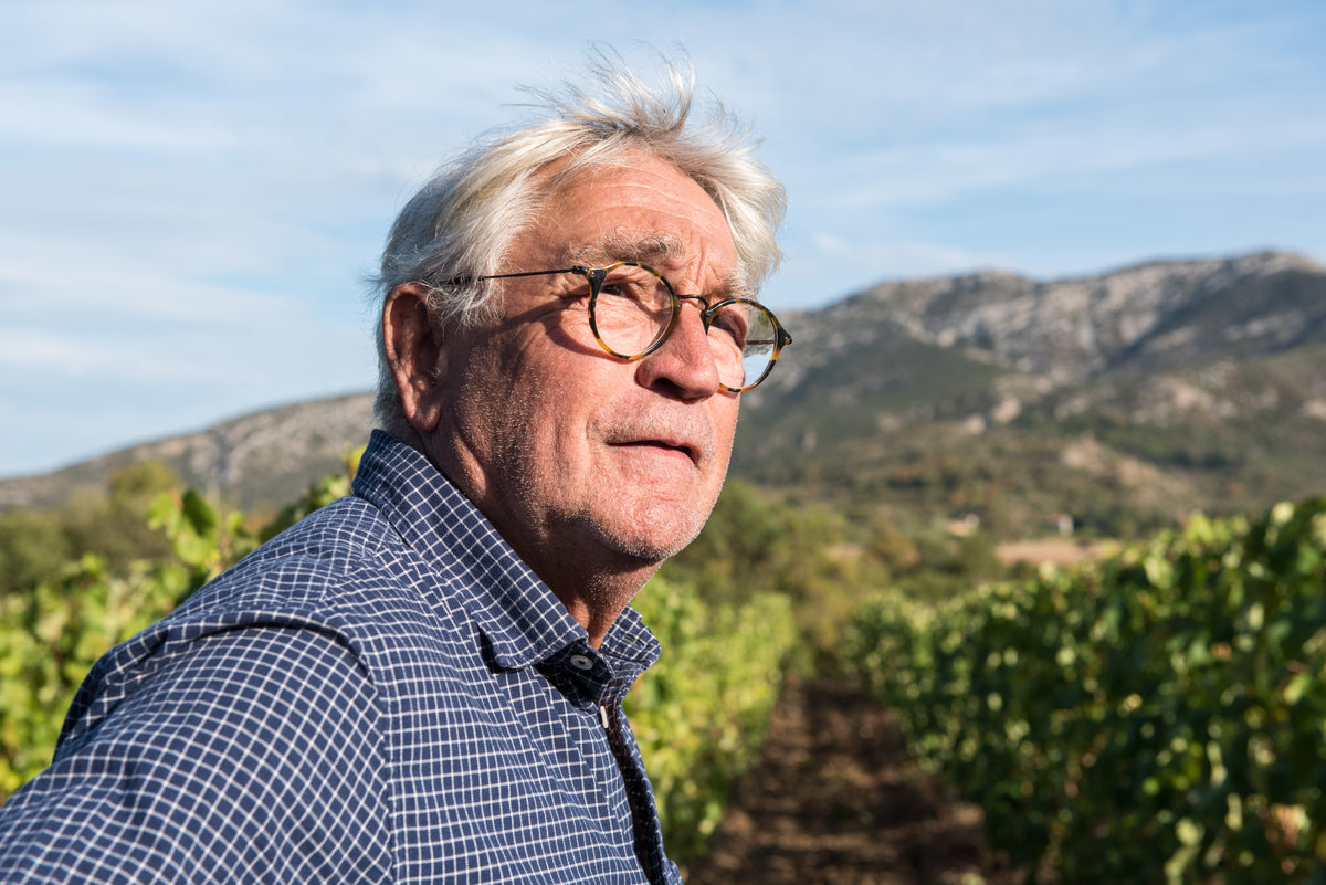 An older man with gray hair and glasses stands in a vineyard, wearing a blue checked shirt. Green vines and distant mountains are visible under a clear sky.