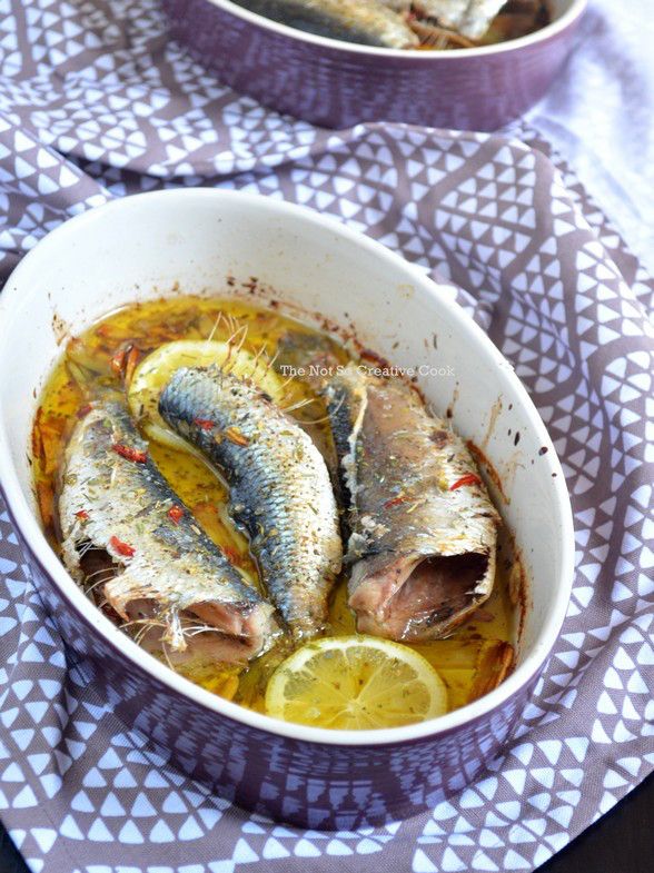Three whole sardines are baked in a white oval dish with lemon slices, herbs, and oil. The dish sits on a patterned cloth, and another similar dish is partially visible in the background.