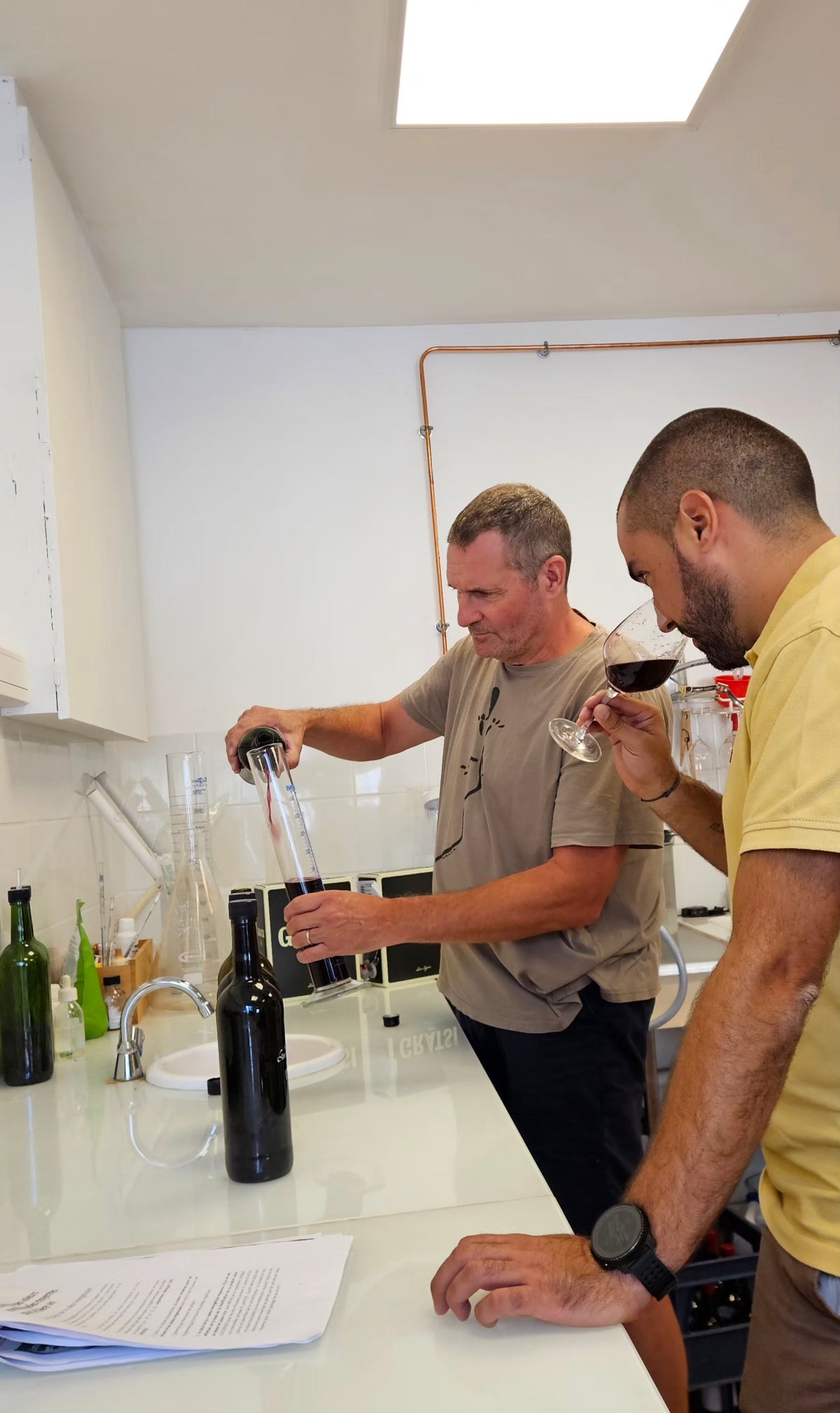Two people in a lab testing wine; one pours wine into a beaker while the other smells a glass. Lab equipment, bottles, and papers are on the counter.