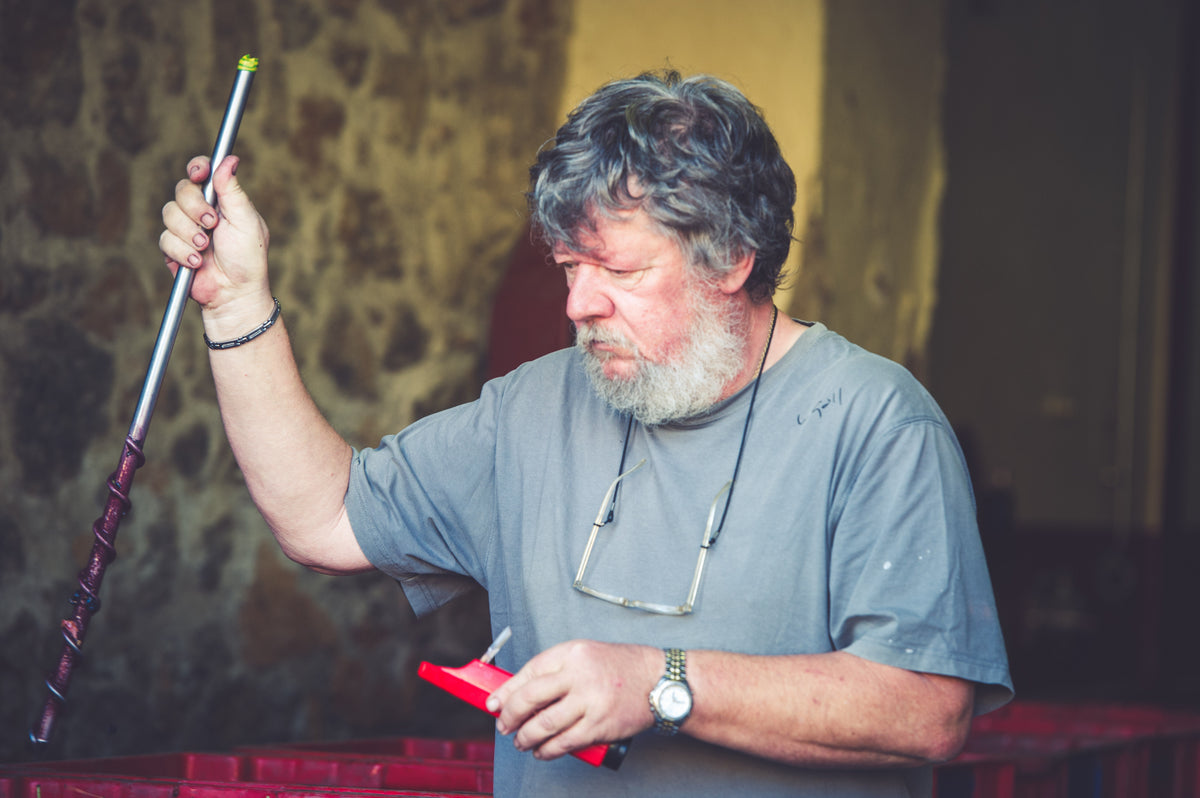 A man with gray hair and a beard, wearing glasses around his neck, holds a metal rod and a red tool, standing indoors near a stone wall.