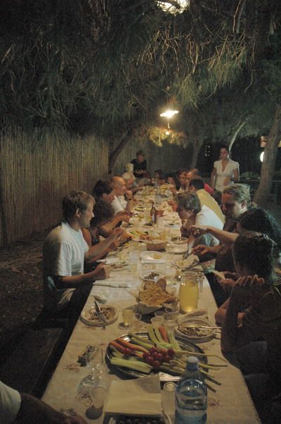 A large group of people sit around a long outdoor table at night, sharing a meal under trees with overhead lights. The table is set with plates, drinks, and various dishes. The atmosphere is lively and communal.