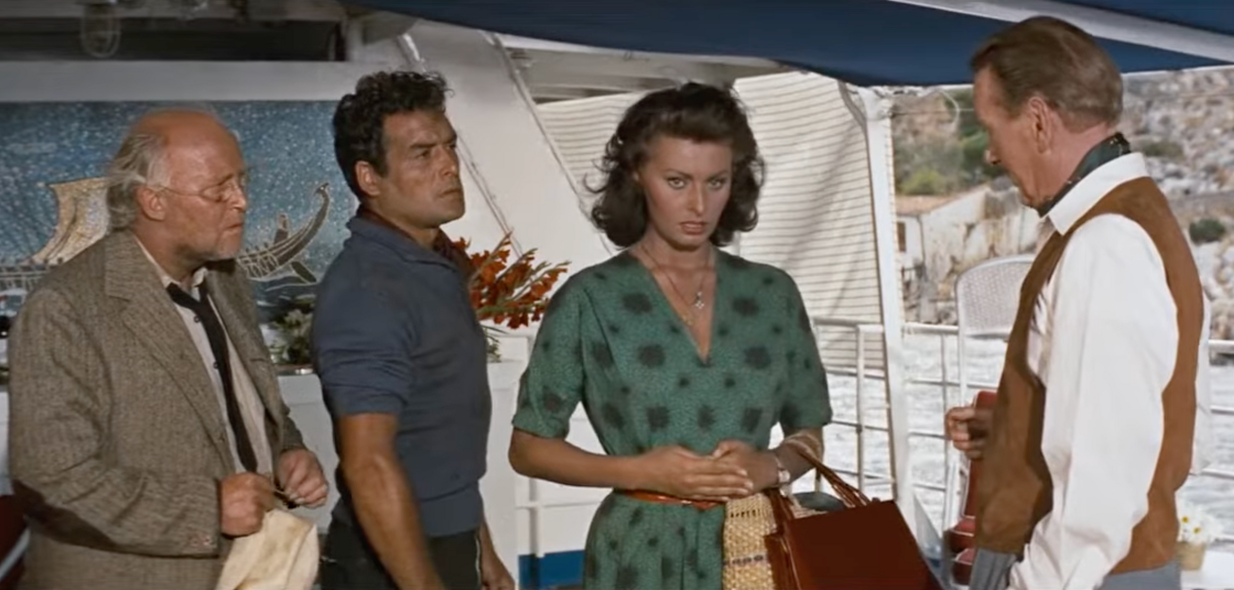 Four people stand on a boat; a woman in a green dress looks forward, flanked by two men. Another man in a vest stands opposite her, holding something. A painting and water are visible in the background.