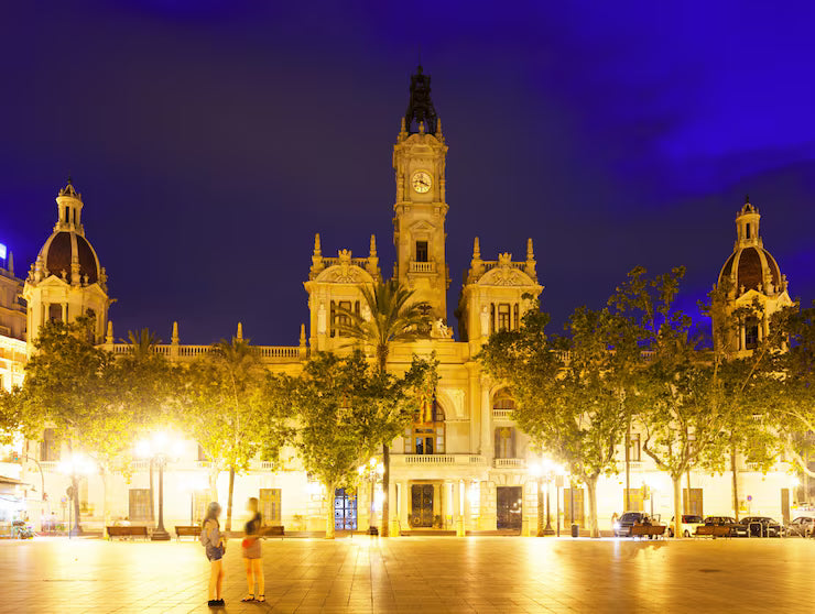 Night view of a grand, illuminated historic building with ornate towers and a clock, set against a deep blue sky, with two people standing in a spacious plaza in front.