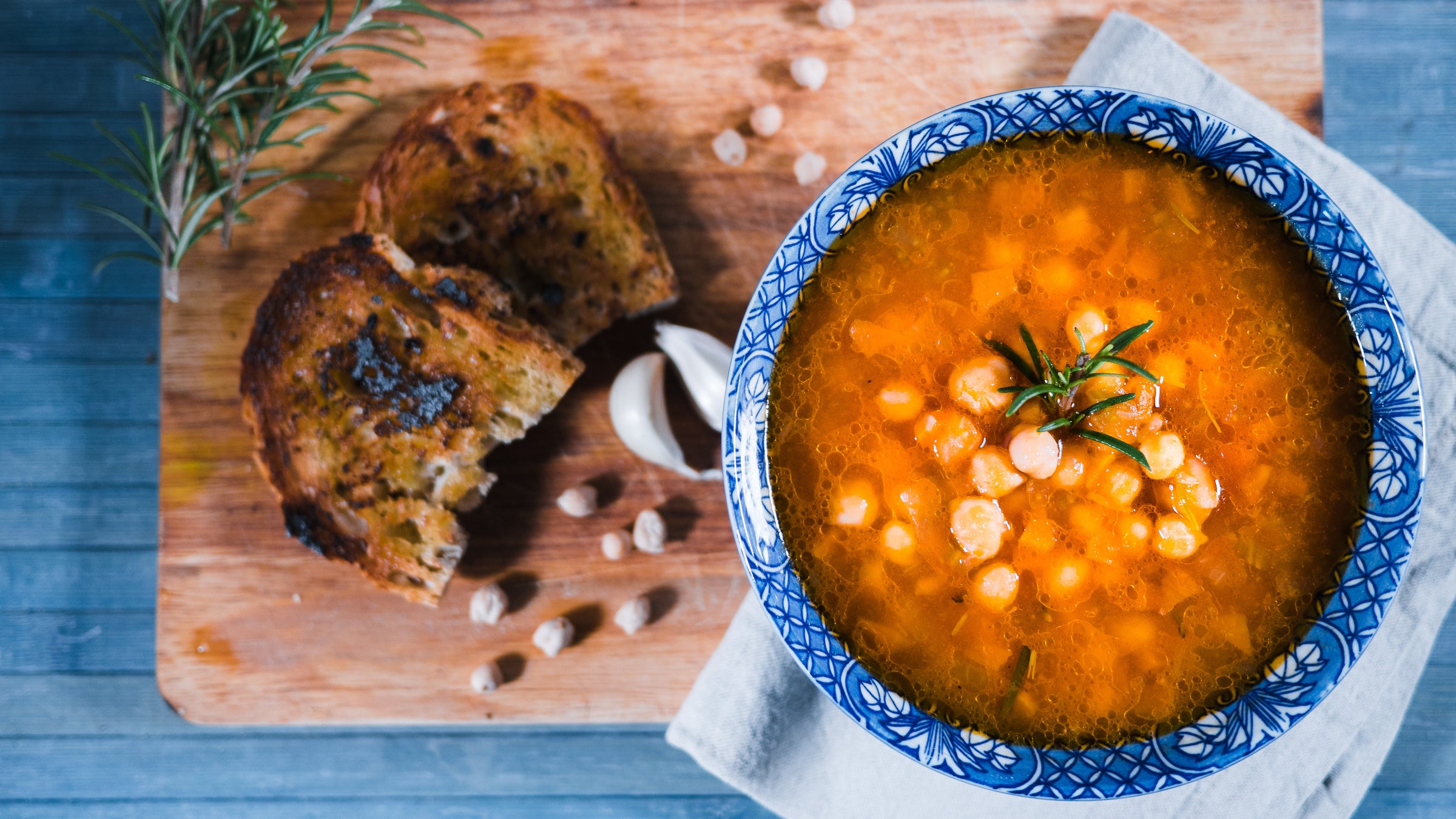 A bowl of chickpea soup with herbs, served on a napkin next to toasted bread, garlic, and loose chickpeas on a wooden board.