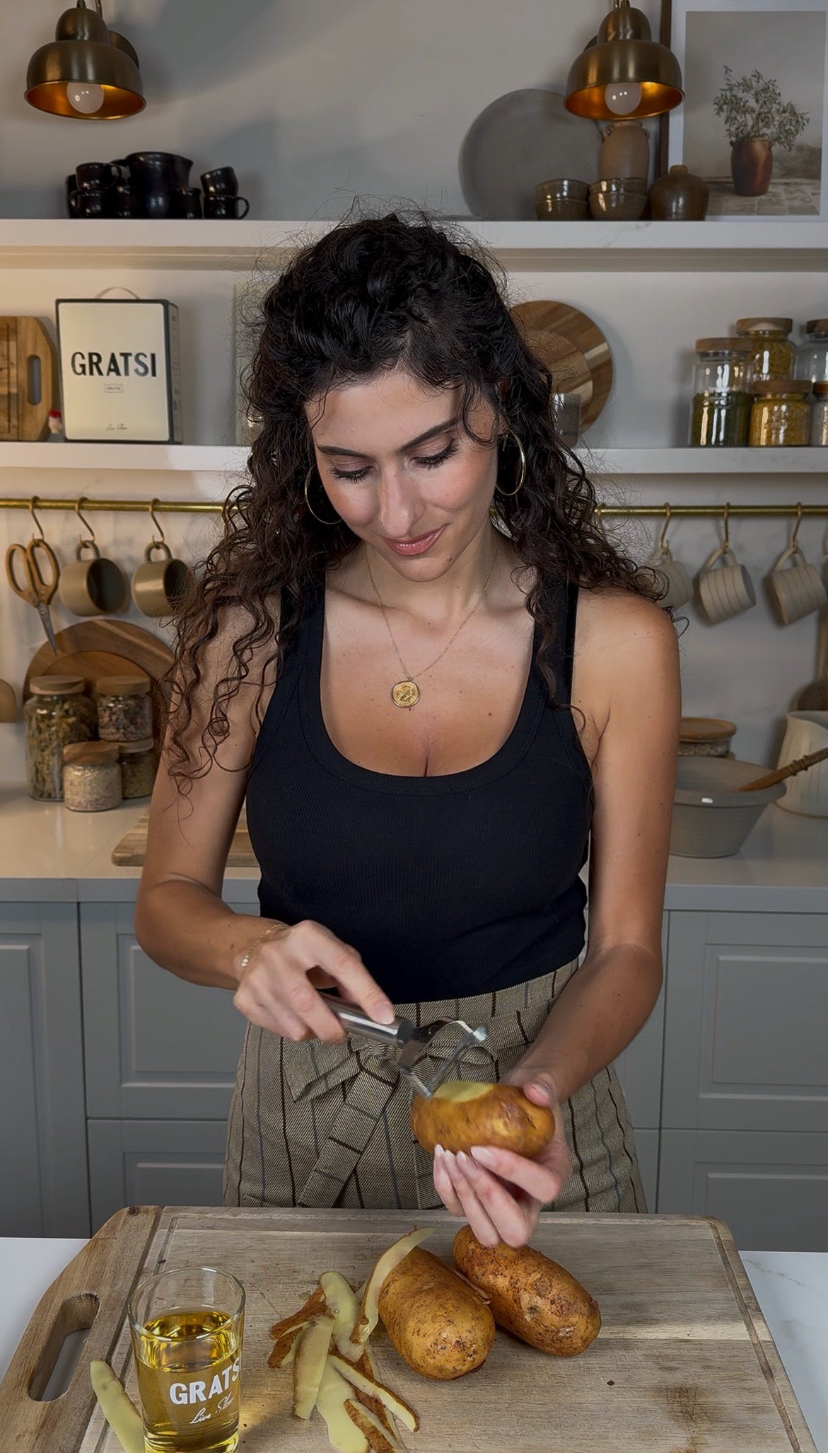 A woman with curly hair wearing a black tank top and striped apron is scooping out a baked potato in a modern kitchen, with kitchen utensils and jars visible in the background.