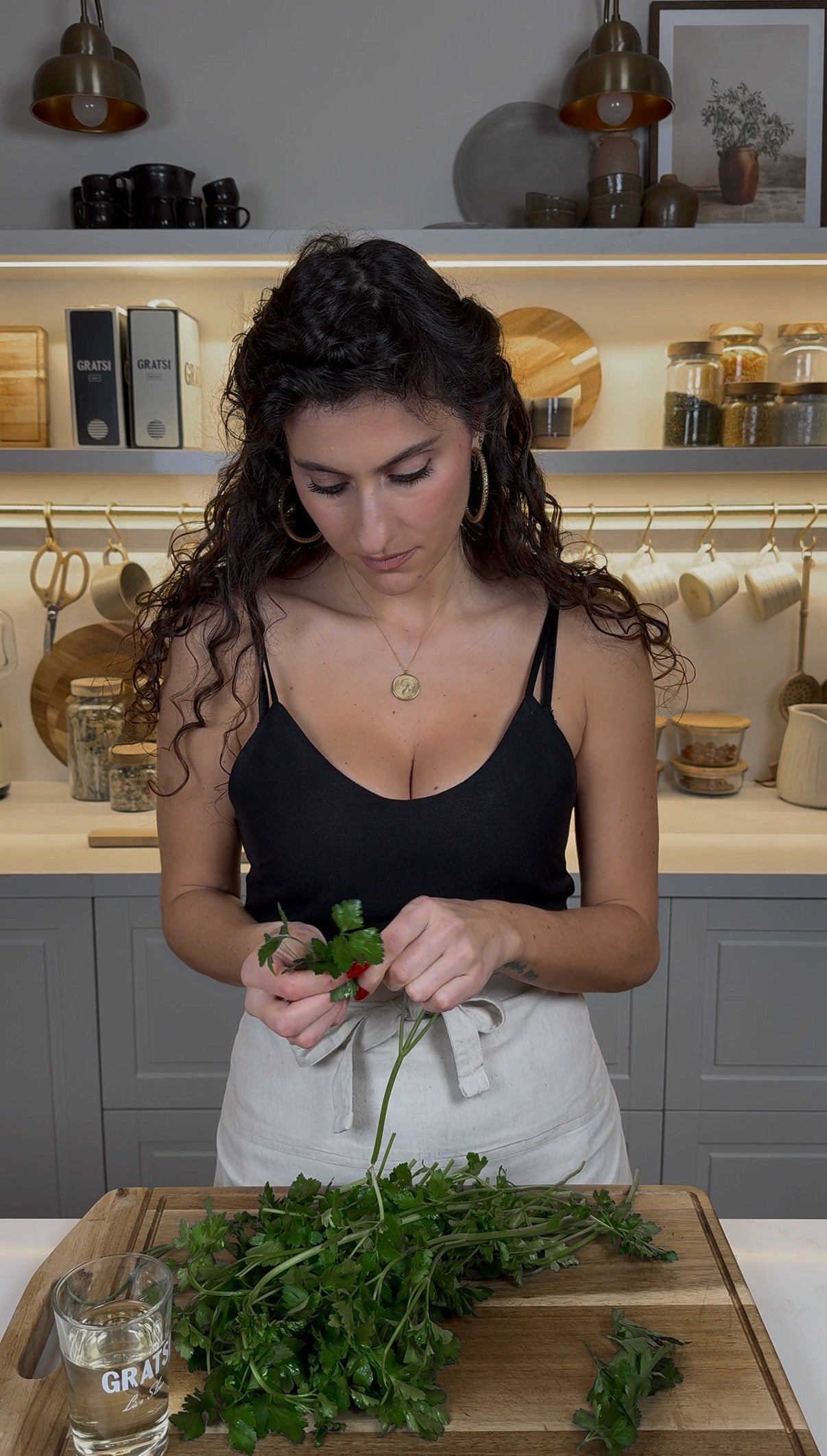 A woman with curly hair wearing a black tank top and white apron stands in a modern kitchen, looking down and picking herbs from a bunch of parsley on a wooden cutting board.