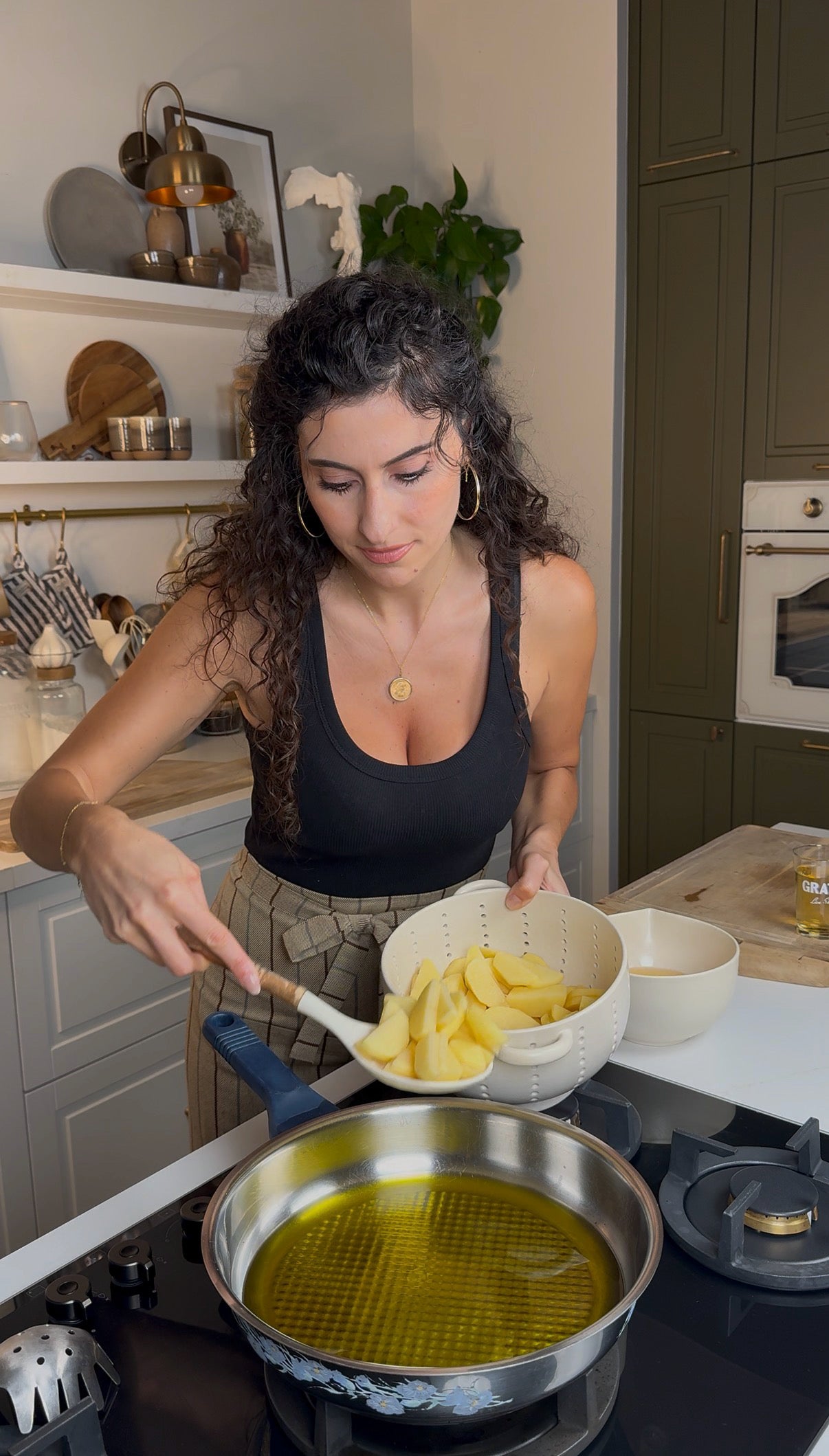 A woman with curly hair and a black tank top adds sliced potatoes from a colander into a frying pan with oil on a stove in a modern kitchen.