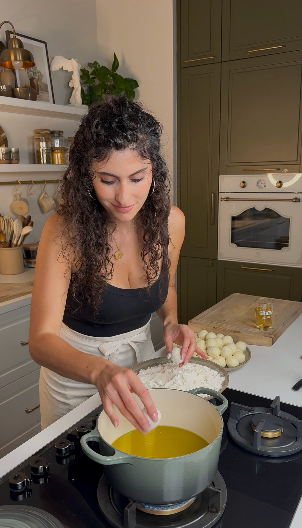 A woman with curly hair and a black tank top prepares food in a kitchen, adding an ingredient to a pot on the stove with a tray of dough balls and flour beside her. Shelves with jars and utensils are in the background.
