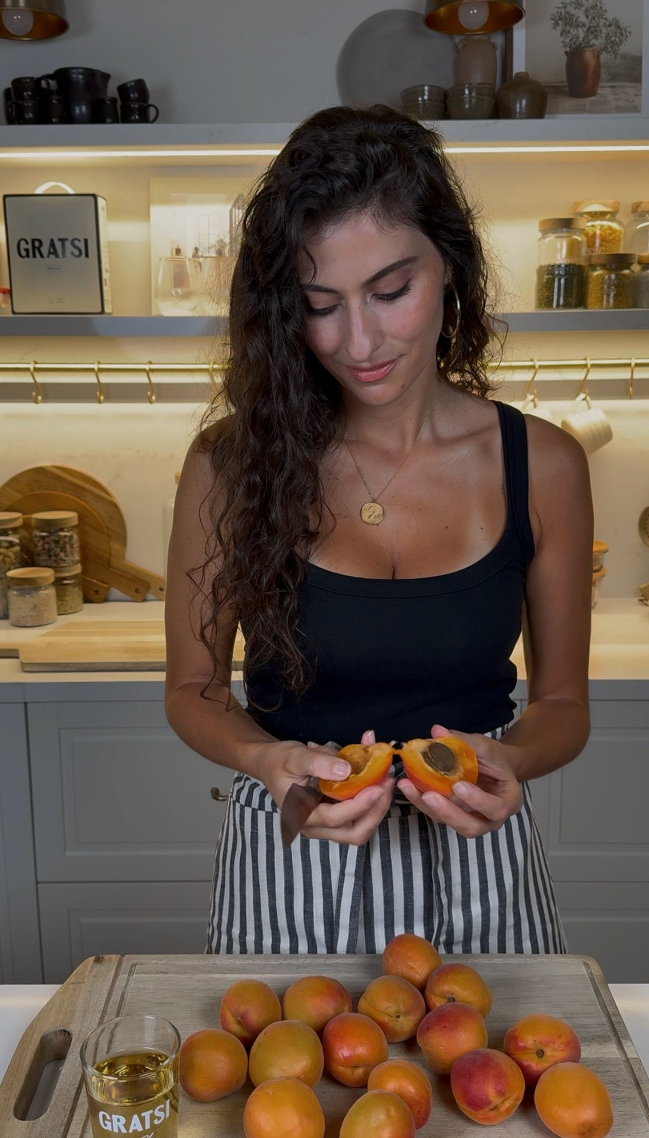Woman with long curly hair in a black tank top and striped apron stands in a kitchen, looking down while splitting an apricot in half. Other apricots are on the counter in front of her. Shelves and jars are in the background.