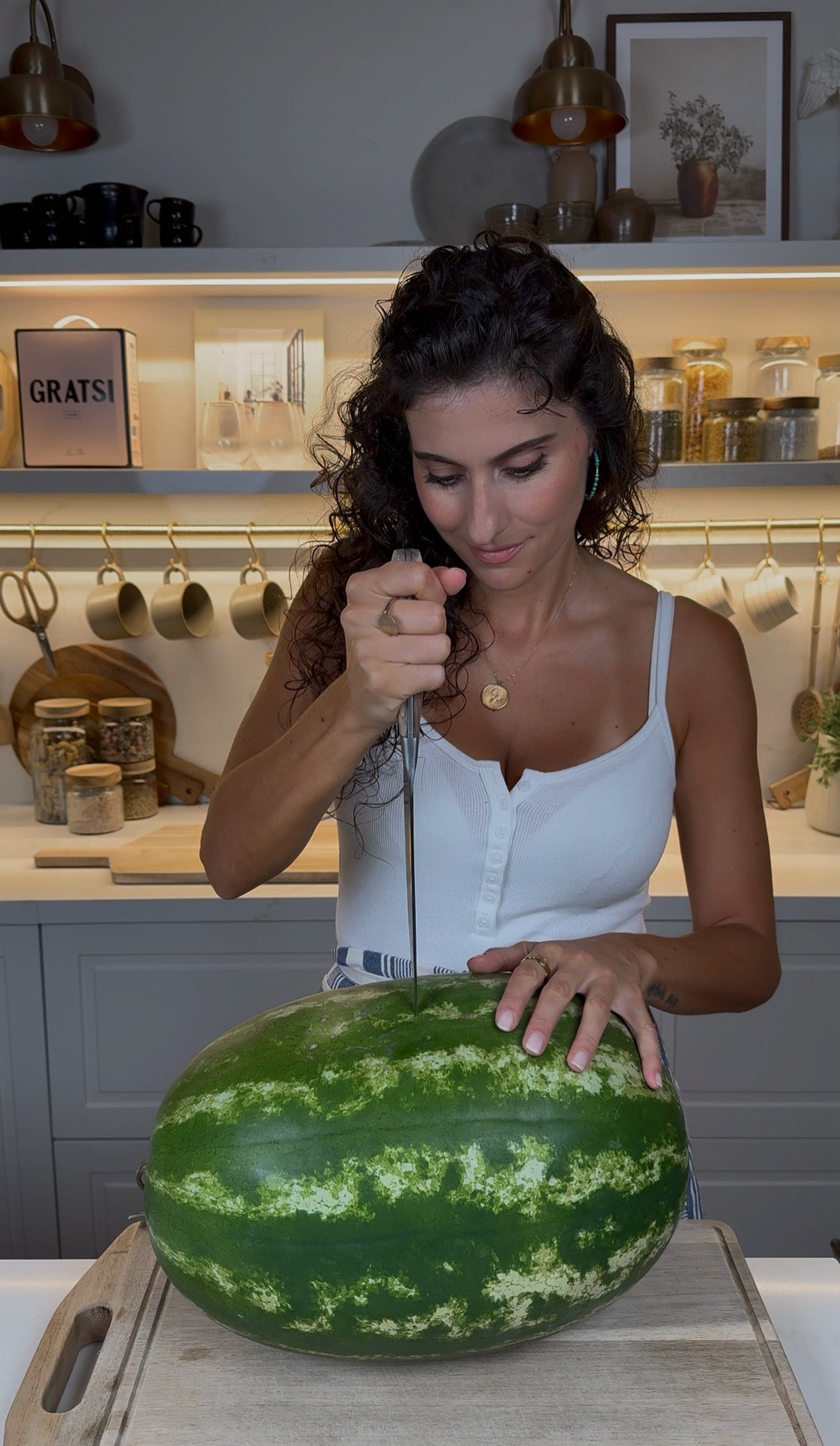 A woman with curly hair in a white top holds a large knife vertically, preparing to cut into a whole watermelon on a kitchen counter. Kitchen shelves and utensils are visible in the background.