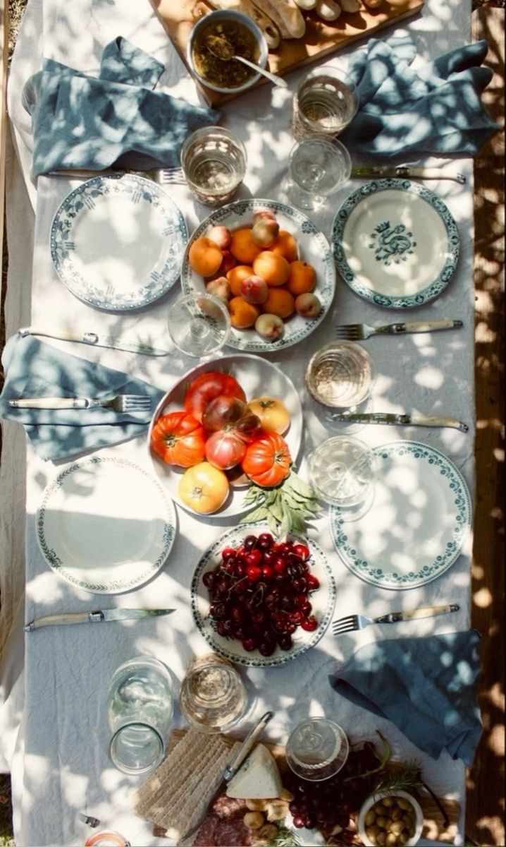 A sunlit outdoor table set for a meal with plates, blue napkins, glasses, and bowls of fresh fruit, including cherries, tomatoes, and apricots, plus olives and cheeses. Dappled shadows from tree leaves fall across the table.