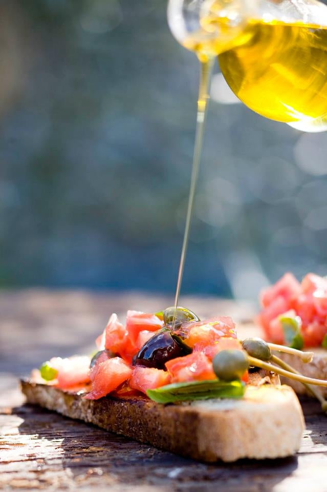 A close-up of bruschetta topped with chopped tomatoes, olives, and capers on rustic bread, with golden olive oil being poured from above.