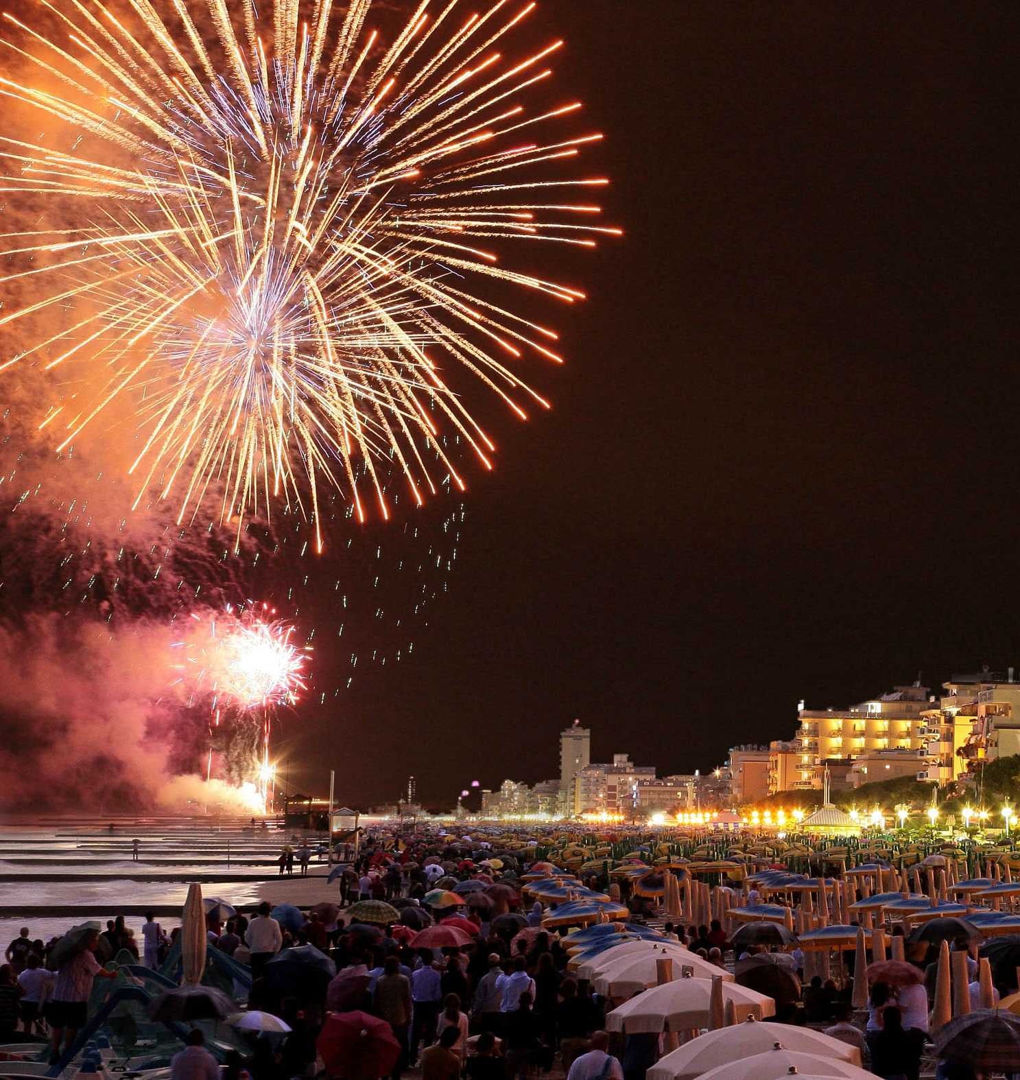 Fireworks light up the night sky over a crowded beach lined with umbrellas and people, with city buildings illuminated in the background.