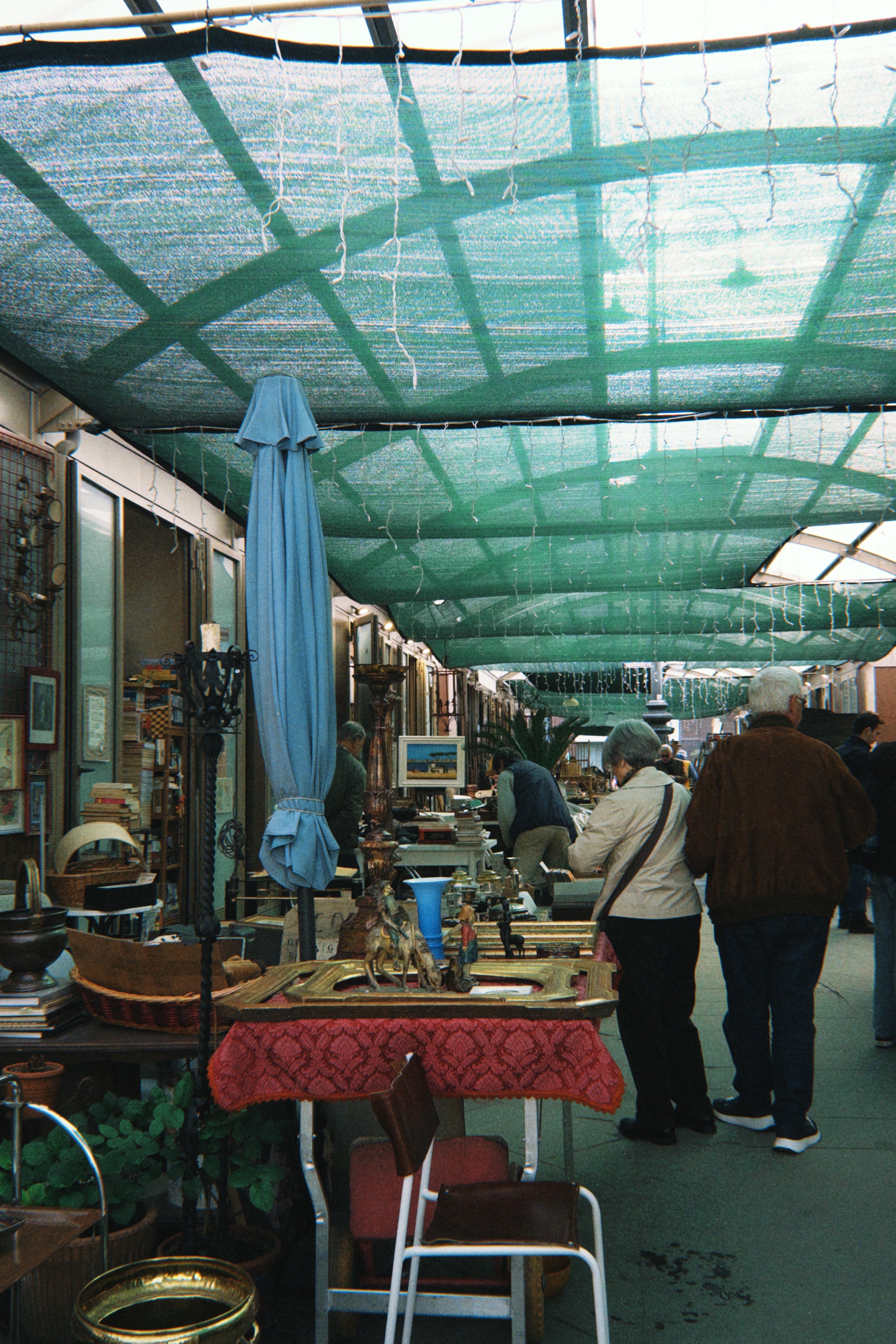 People browse various vintage and antique items displayed on tables at an outdoor market under a green canopy. A red tablecloth, blue closed umbrella, and framed art are visible in the foreground.