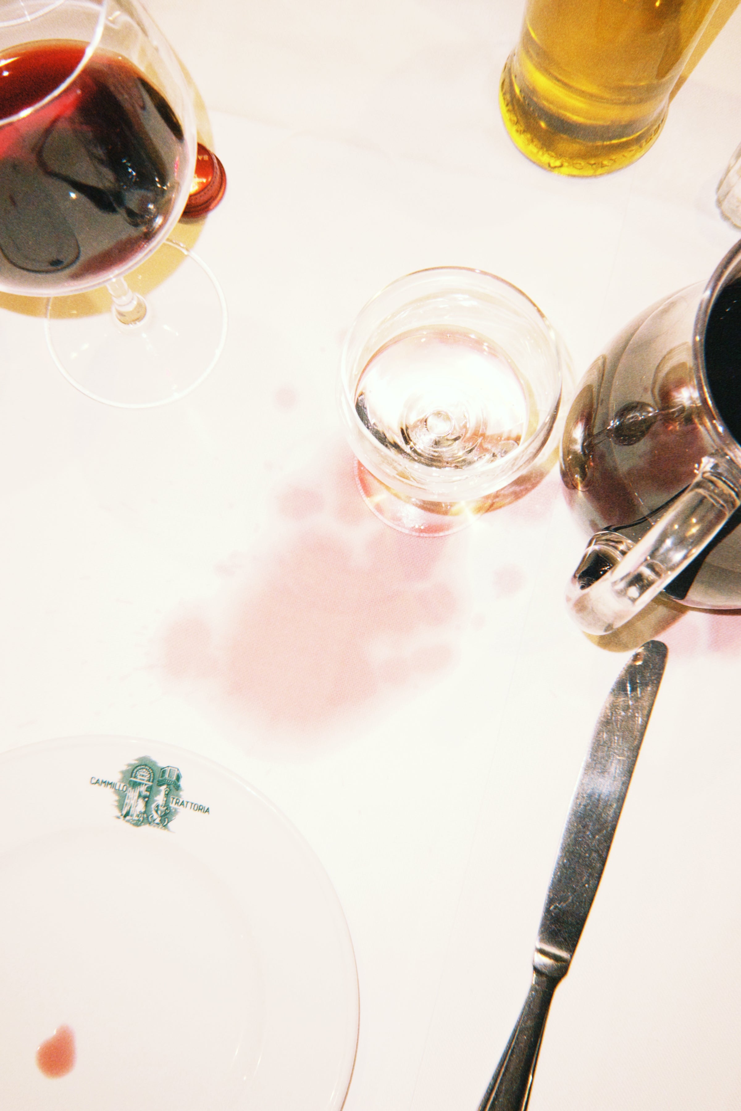 A glass of wine has spilled on a white tablecloth, surrounded by a wine glass, a water glass, a pitcher, a knife, and a plate with a small food stain.