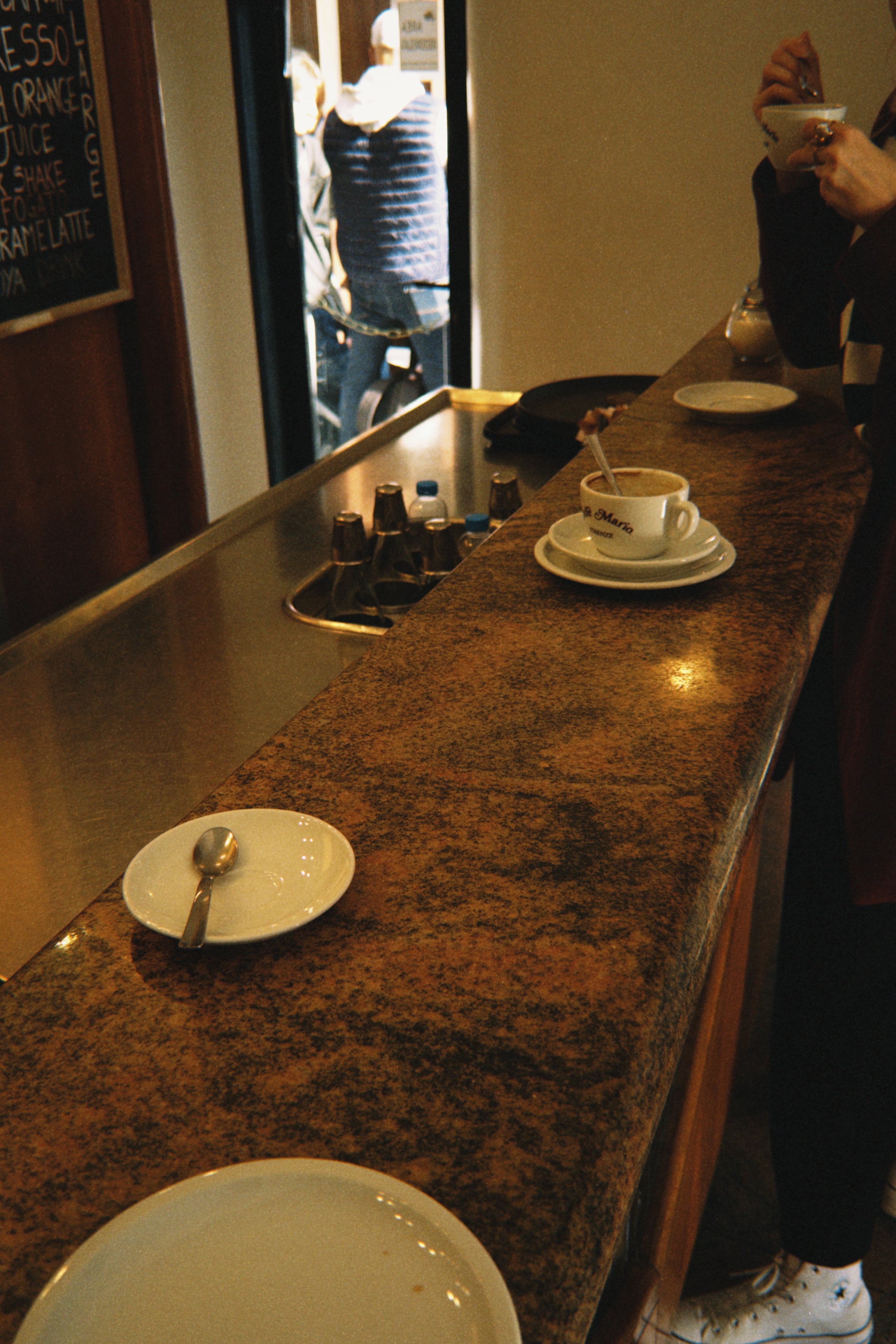 A granite coffee bar with a cup and saucer in the foreground, empty plates with spoons, and a person holding a cup. Another person is partially visible outside through the door.