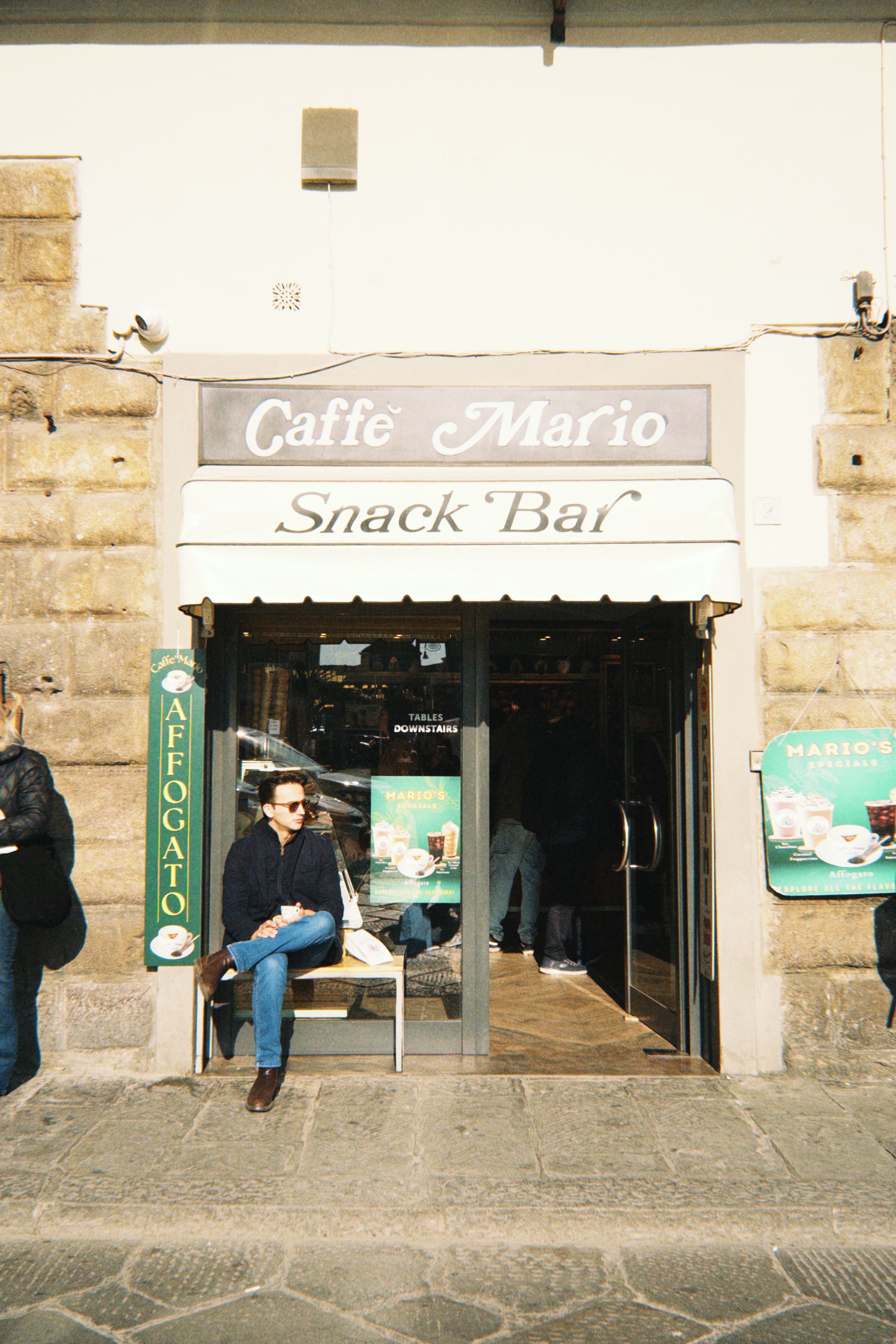 A man in sunglasses sits on a bench outside Caffè Mario Snack Bar, with stone walls and signs nearby. The entrance is open and other people are walking past.