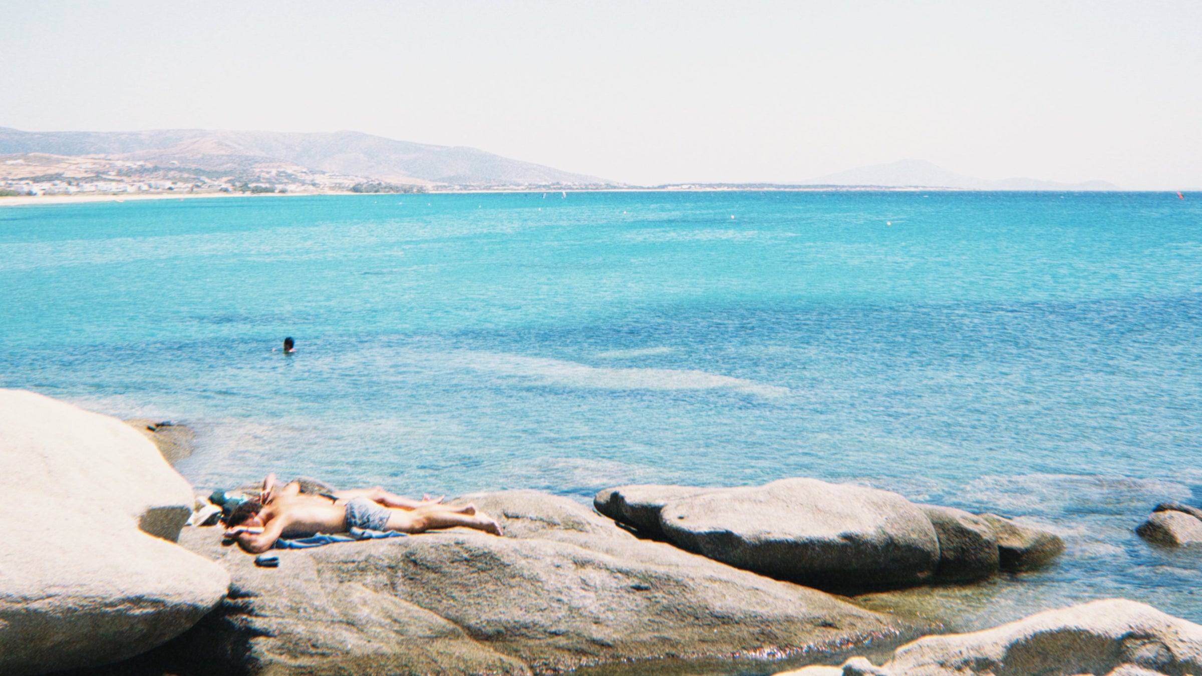 A person sunbathes on large coastal rocks beside clear blue sea water, while another person swims nearby; mountains and a distant shoreline are visible in the background under a clear sky.