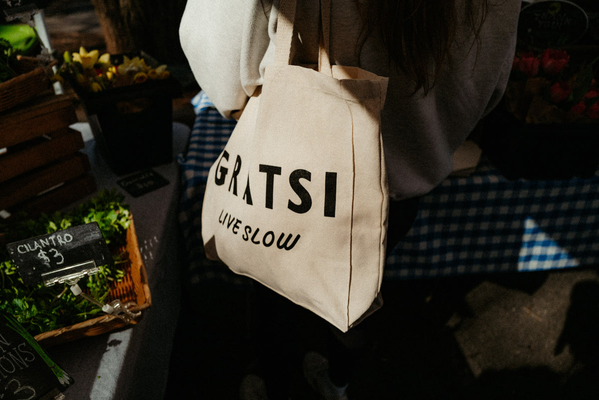 A person carries a beige tote bag reading GRATSI LIVE SLOW at an outdoor market, with herbs and flowers for sale nearby.