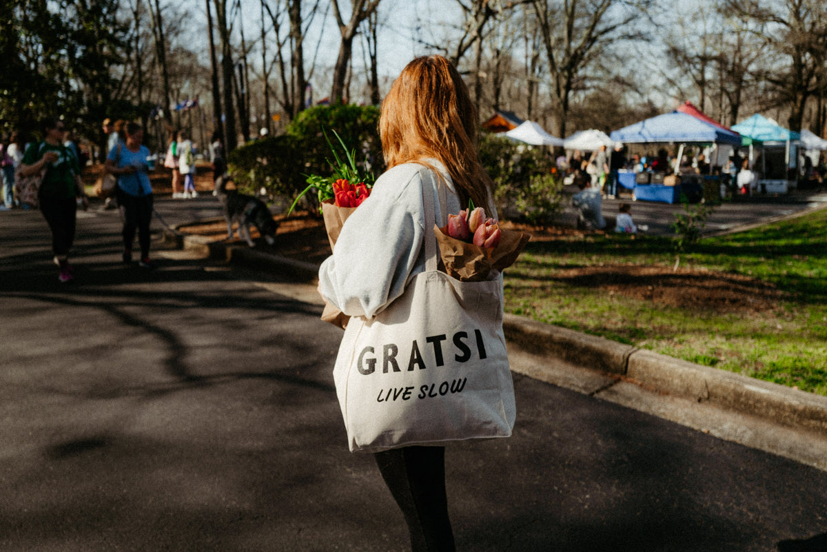 A person with long hair carries flowers in a reusable GRATSI LIVE SLOW tote at an outdoor market. Tents, trees, and other people are visible in the background.