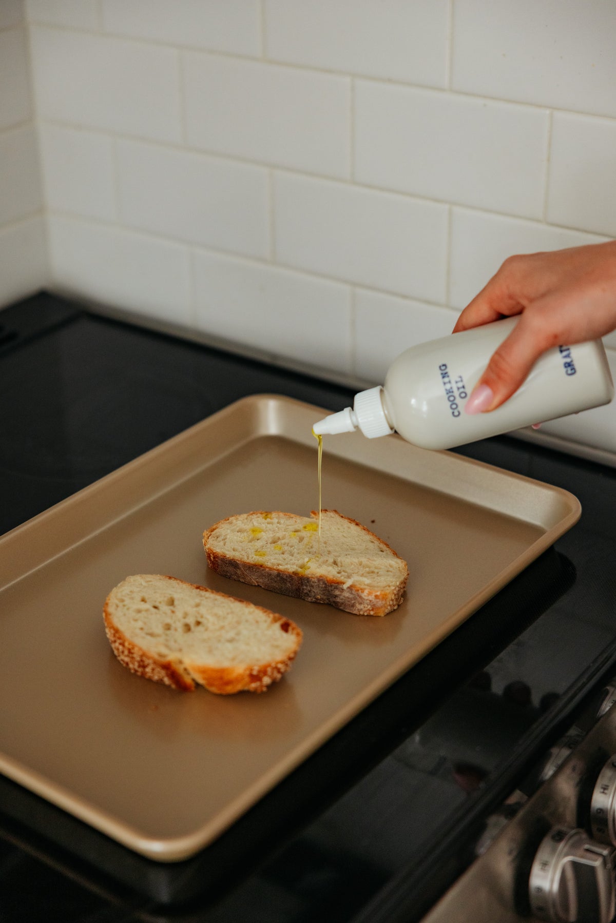 A hand squeezes a bottle of cooking oil, drizzling it onto slices of bread on a baking sheet.