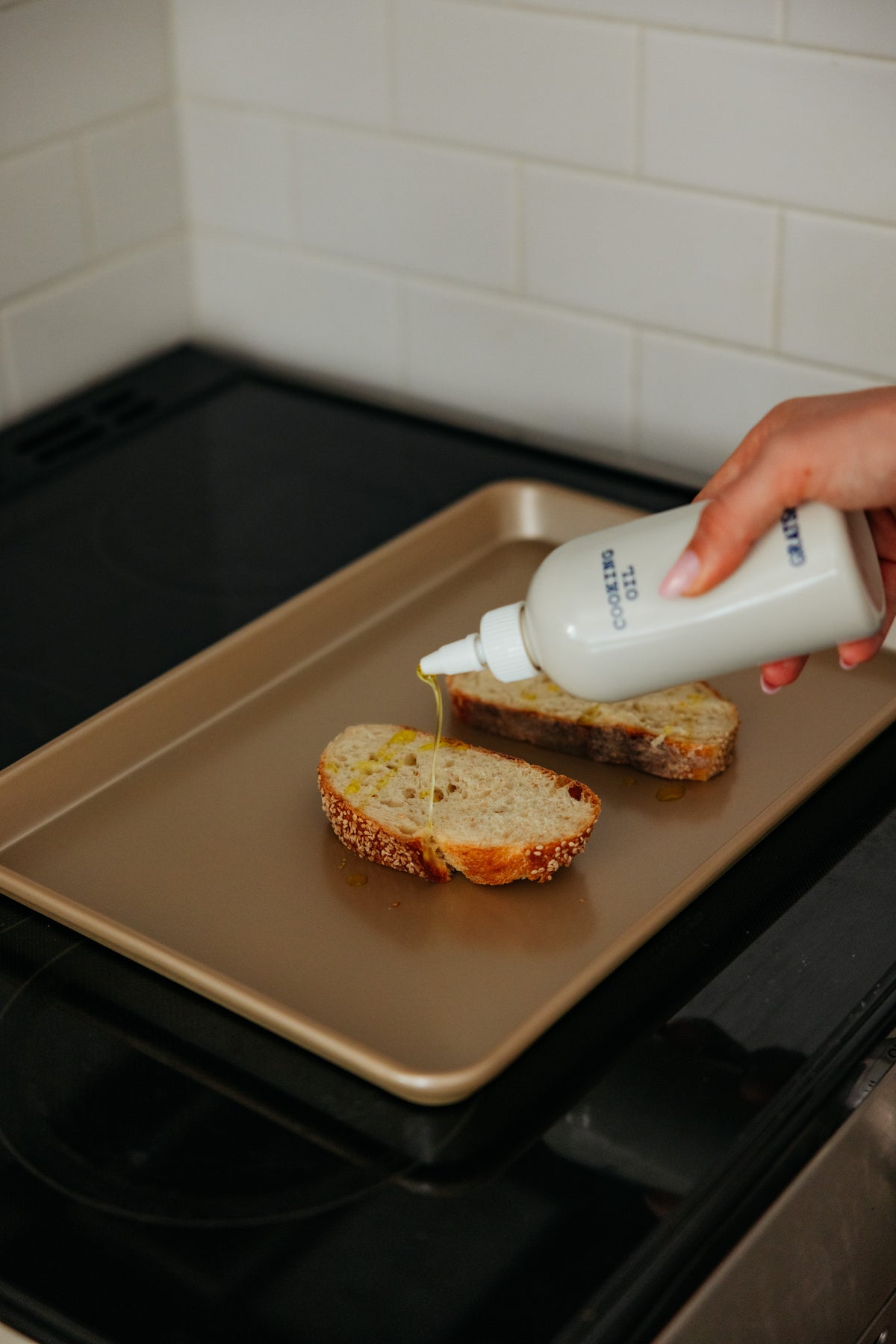 A hand squeezes a bottle of cooking oil onto two slices of bread on a baking sheet.