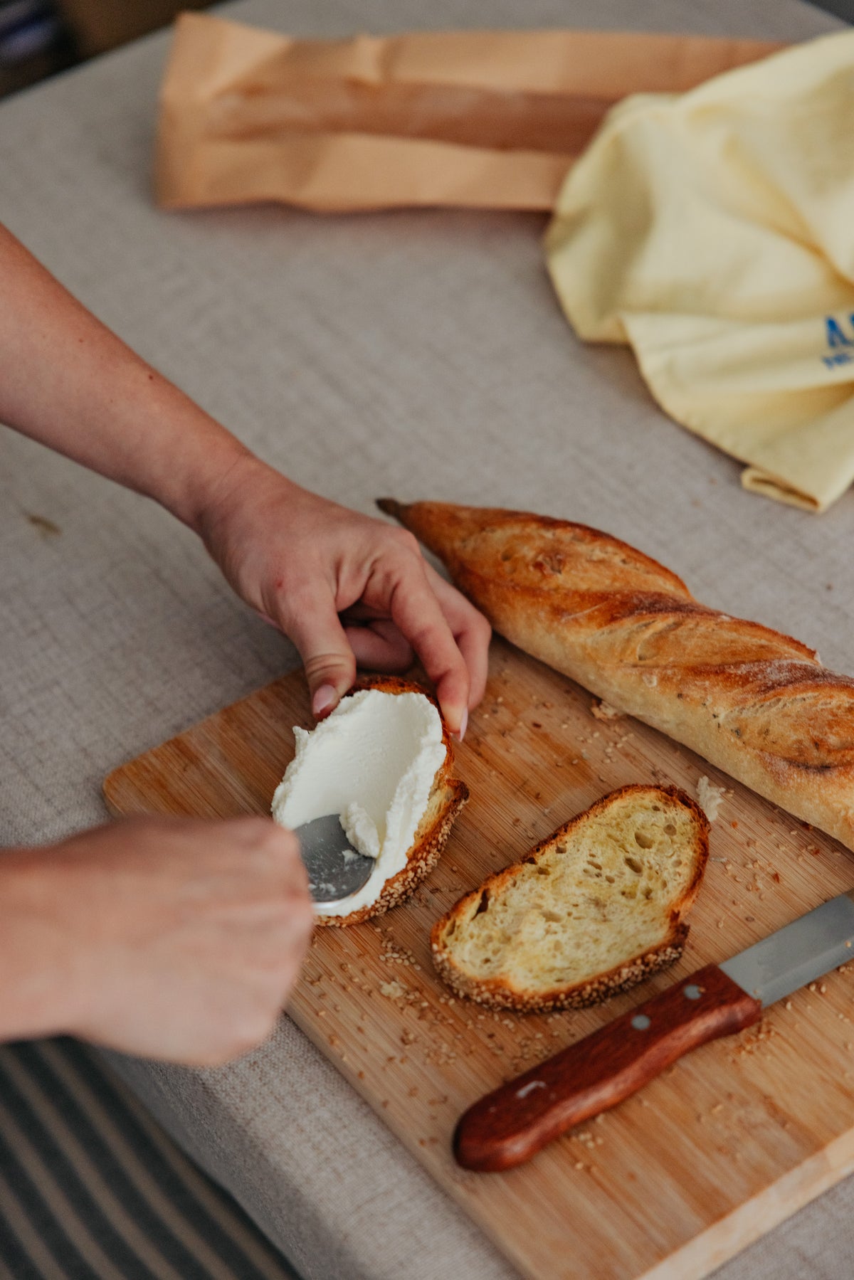 A person uses a knife to spread cream cheese onto a slice of bread on a wooden cutting board, with a baguette nearby.