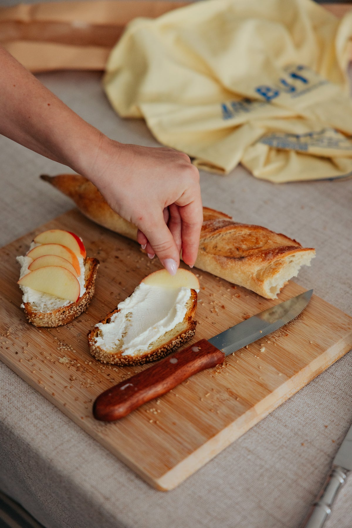A hand places a thin apple slice onto a piece of bread spread with cream cheese, next to a baguette and a knife on a wooden cutting board.