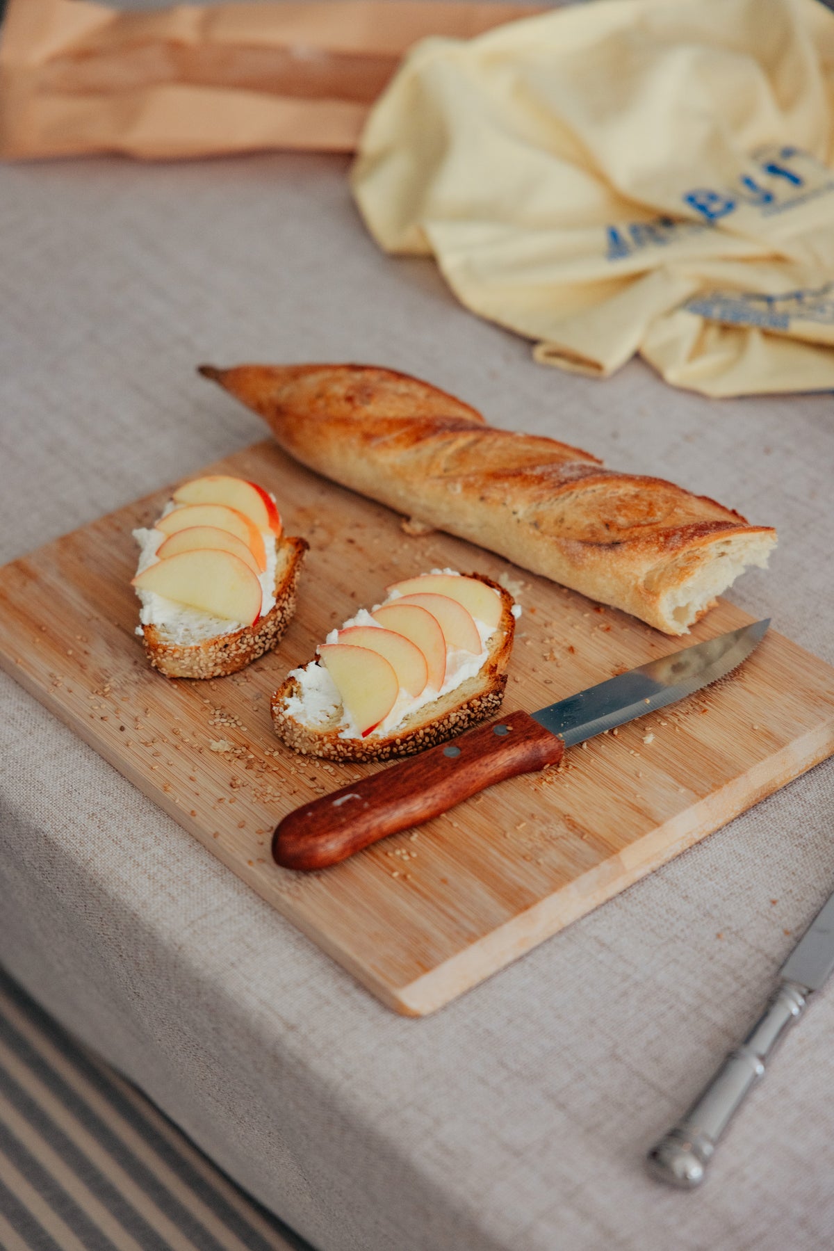 Two slices of bread topped with cream cheese and apple slices, a baguette, and a knife on a wooden cutting board.