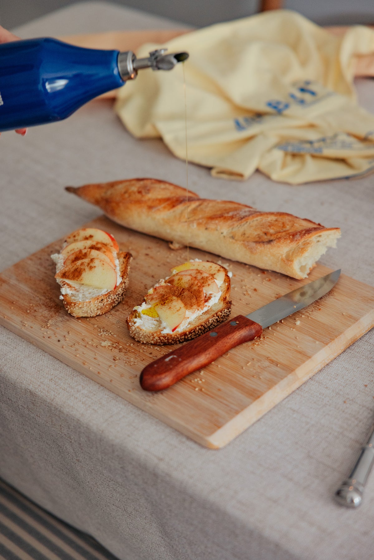 Olive oil being poured from a blue bottle onto two slices of bread topped with cream cheese, apple slices, and cinnamon, next to a baguette and a knife on a wooden board.