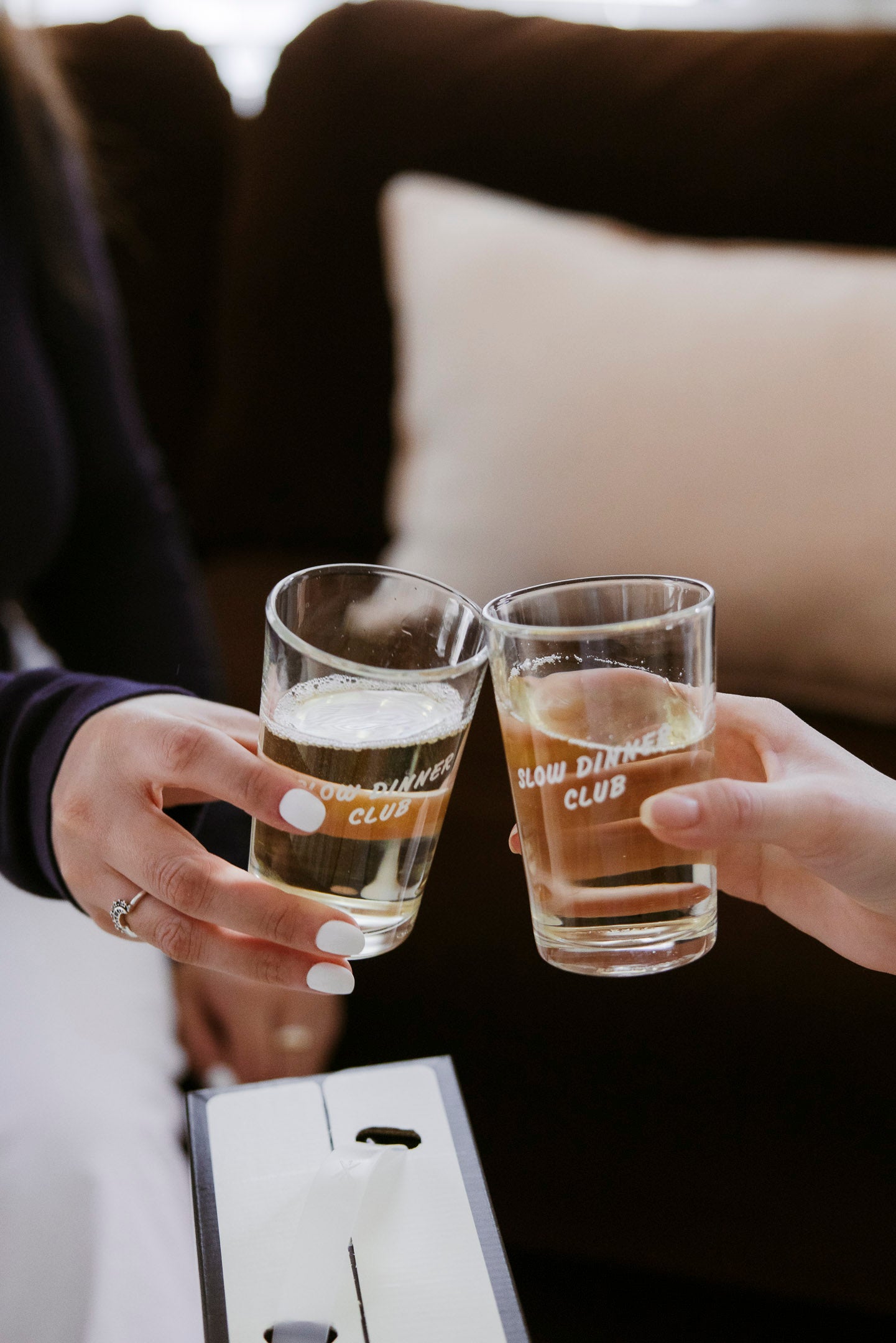 Two people clink glasses of light-colored drinks with ice. The glasses have SLOW DINNER CLUB printed on them. One person wears a ring and has white-painted nails. A couch and cushion are visible in the background.