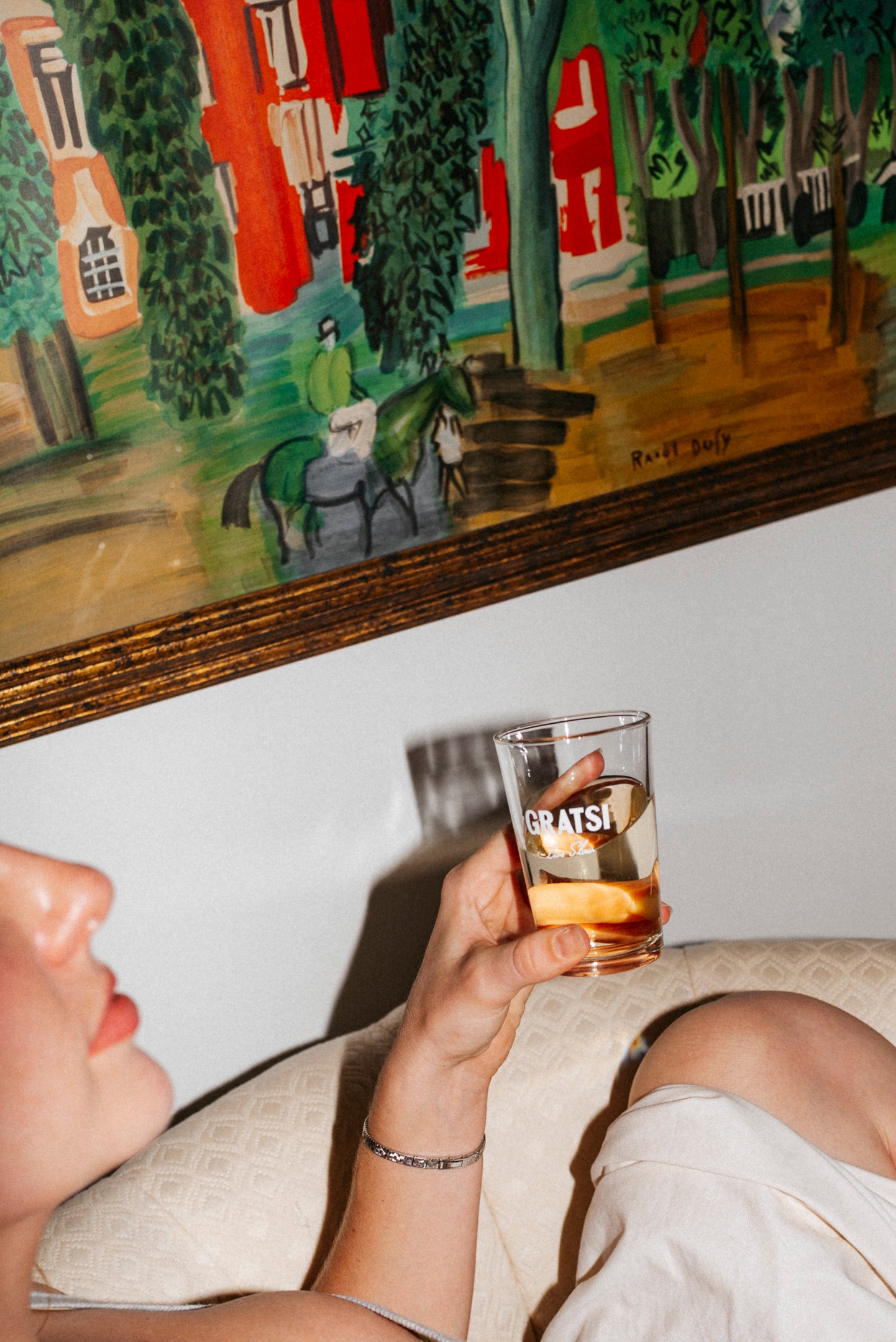 A person sitting on a light-colored couch holds a glass of amber liquid with ice that says GRATSI. In the background, there is a colorful painting of a person riding a horse.