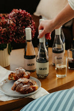 A hand reaches for a Gratsi Wine & Pasta Holiday Bundle on a table with several wine bottles, a glass, croissants, and a vase of hydrangeas in the background.