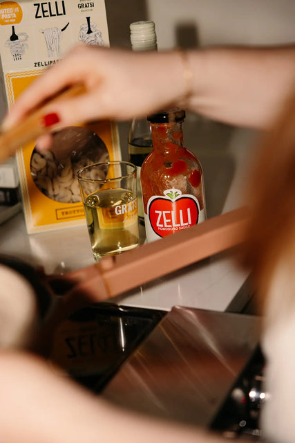 A person cooks near the Gratsi Wine & Pasta Holiday Bundle on the kitchen counter, with their arm and hand visible in the foreground.