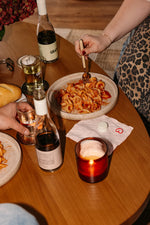 At a wooden table set with wine, glasses, a candle, bread, and a napkin with a red heart, someone in leopard print enjoys pasta using Gratsi’s Wine & Pasta Holiday Bundle while another hand raises a glass beside the plate.