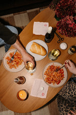 Two people enjoy Gratsi’s Wine & Pasta Holiday Bundle at a wooden table set with pasta in red sauce, white wine, bread, candles, flowers, and festive heart napkins. One reaches for a glass while the other holds a fork.