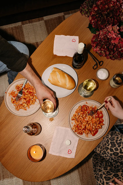 Two people enjoy Gratsi’s Wine & Pasta Holiday Bundle at a wooden table set with pasta in red sauce, white wine, bread, candles, flowers, and festive heart napkins. One reaches for a glass while the other holds a fork.